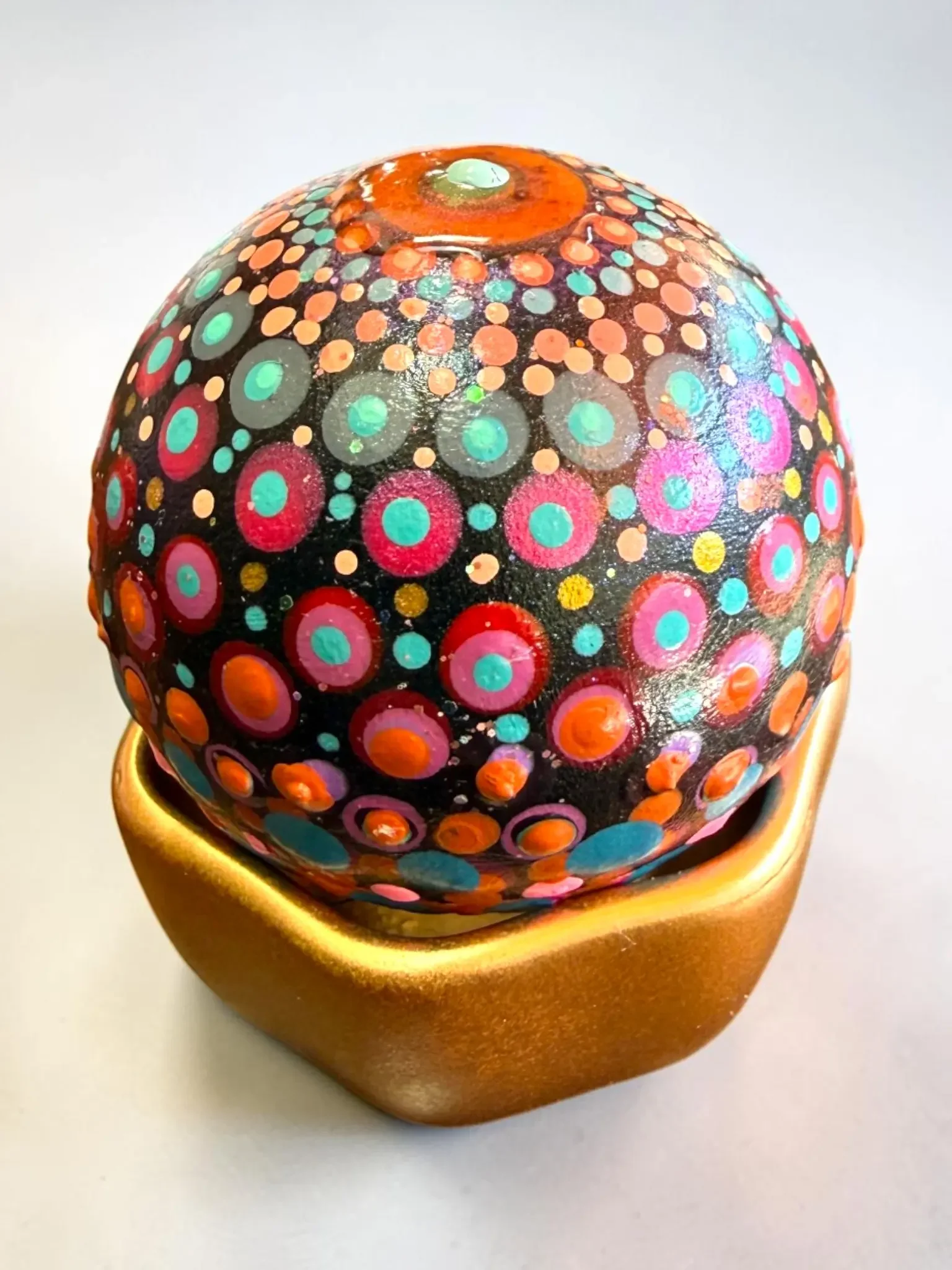 Small globe mandala stone by Color Cult with red, orange, turquoise and pink dot mandala artwork and sits in a golden hand-painted cup.