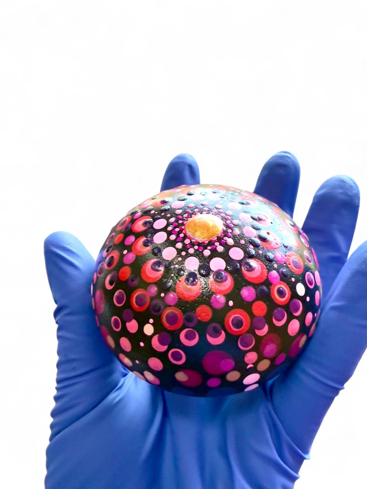 A hand wearing a blue glove holding a black spherical object decorated with colorful, circular dot patterns in pink, purple, and white, with a central yellow dot.