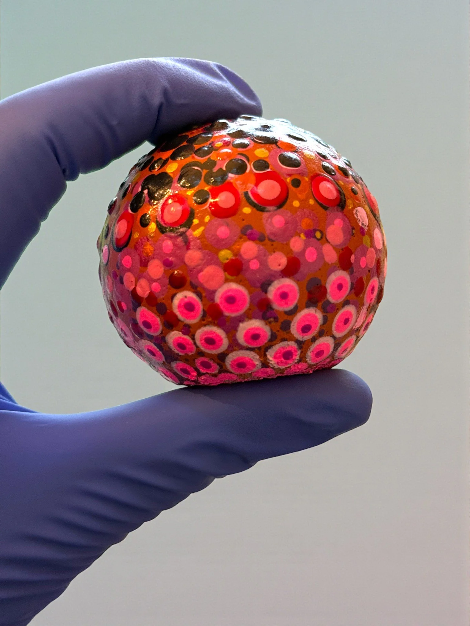 Colorful, decorated object with pink and black circular patterns being held by a hand wearing a purple glove.