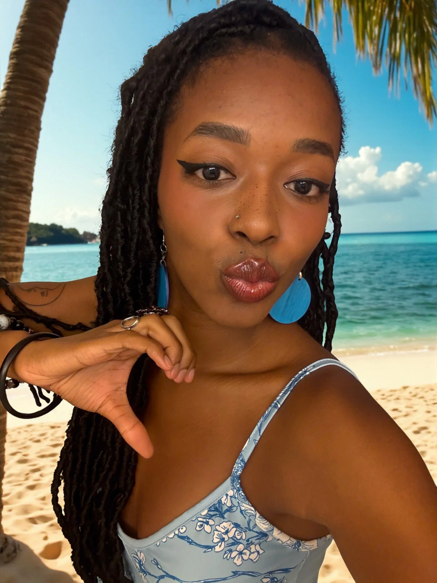 A young woman with dark braided hair, wearing blue earrings and a floral dress, takes a selfie on a tropical beach with palm trees, sand, and the ocean in the background.