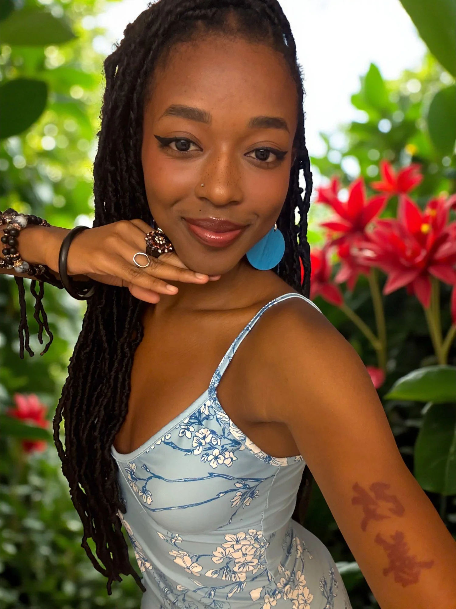 A young woman taking a selfie in front of lush green foliage and red flowers. She has long, braided hair, wears a blue floral dress, large blue earring, and has a tattoo on her right arm.