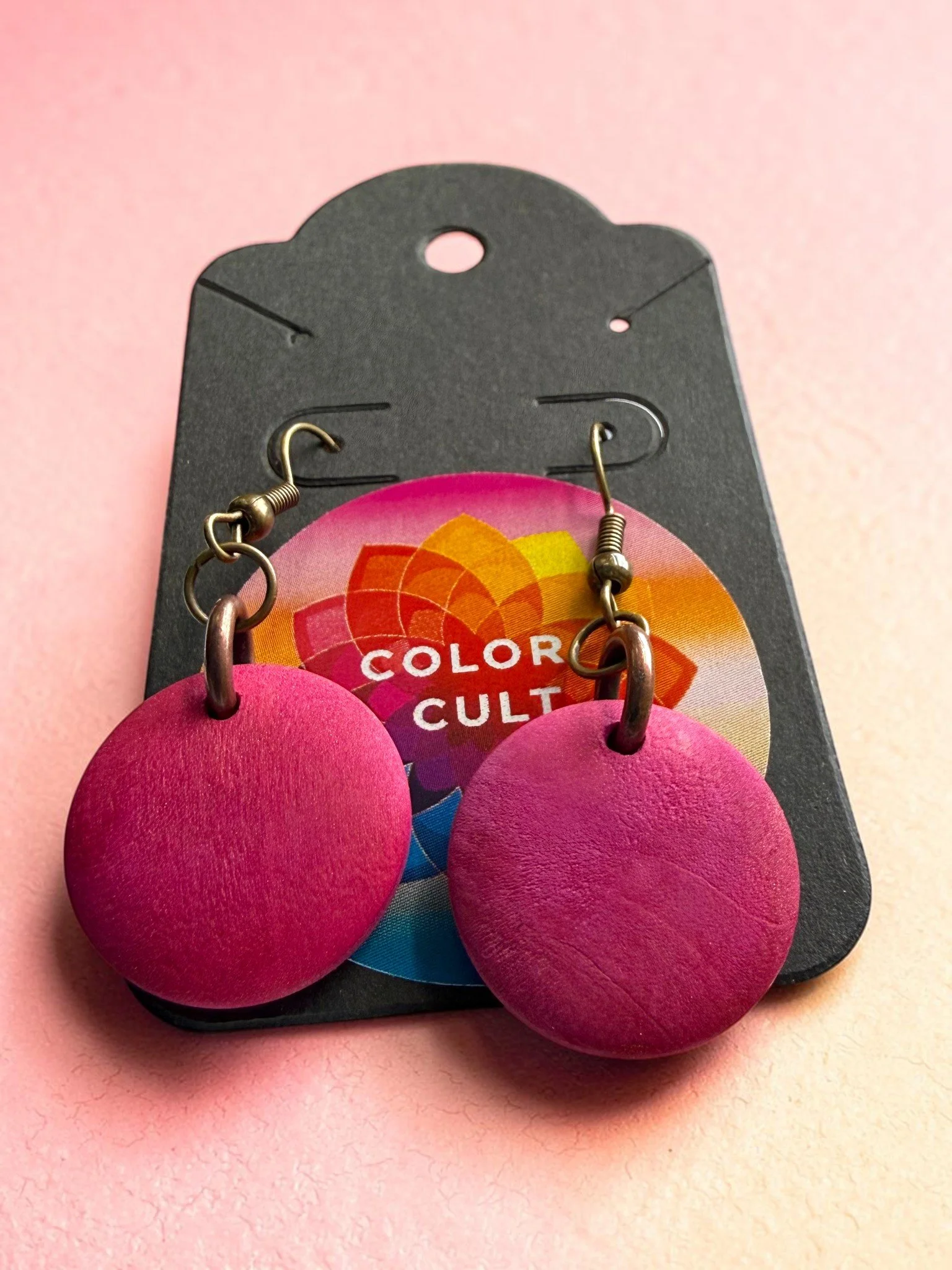 A pair of round, pink earrings with a textured surface, displayed on a black card with colorful graphics and the words "Color Cult."