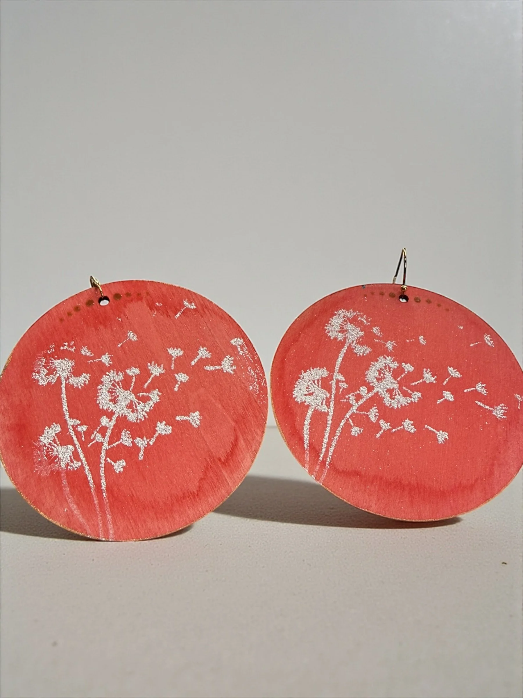 Pair of round coral-red earrings with white floral designs, hanging on a white surface.