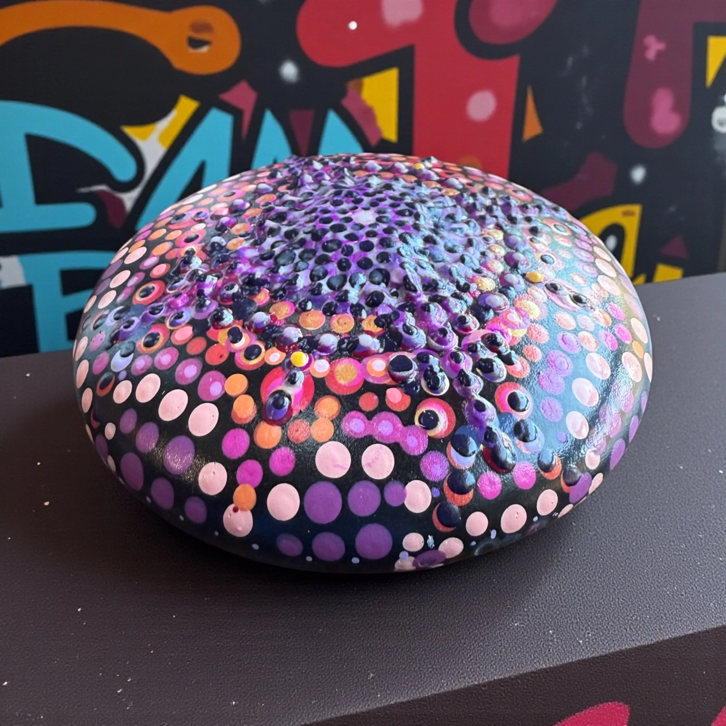 Painted rock with a colorful dot pattern in shades of purple, pink, orange, and black, placed on a black surface with a graffiti-style wall in the background.
