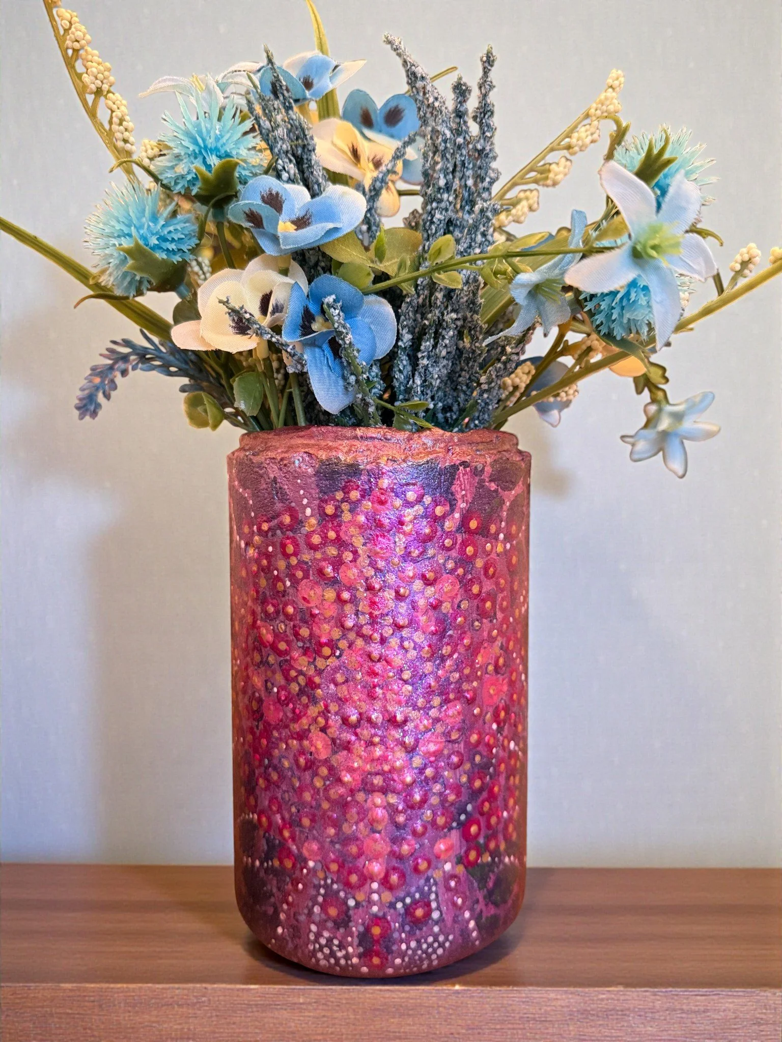 Colorful artificial floral arrangement with blue and yellow flowers in a decorated pink and purple textured vase against a light blue background.