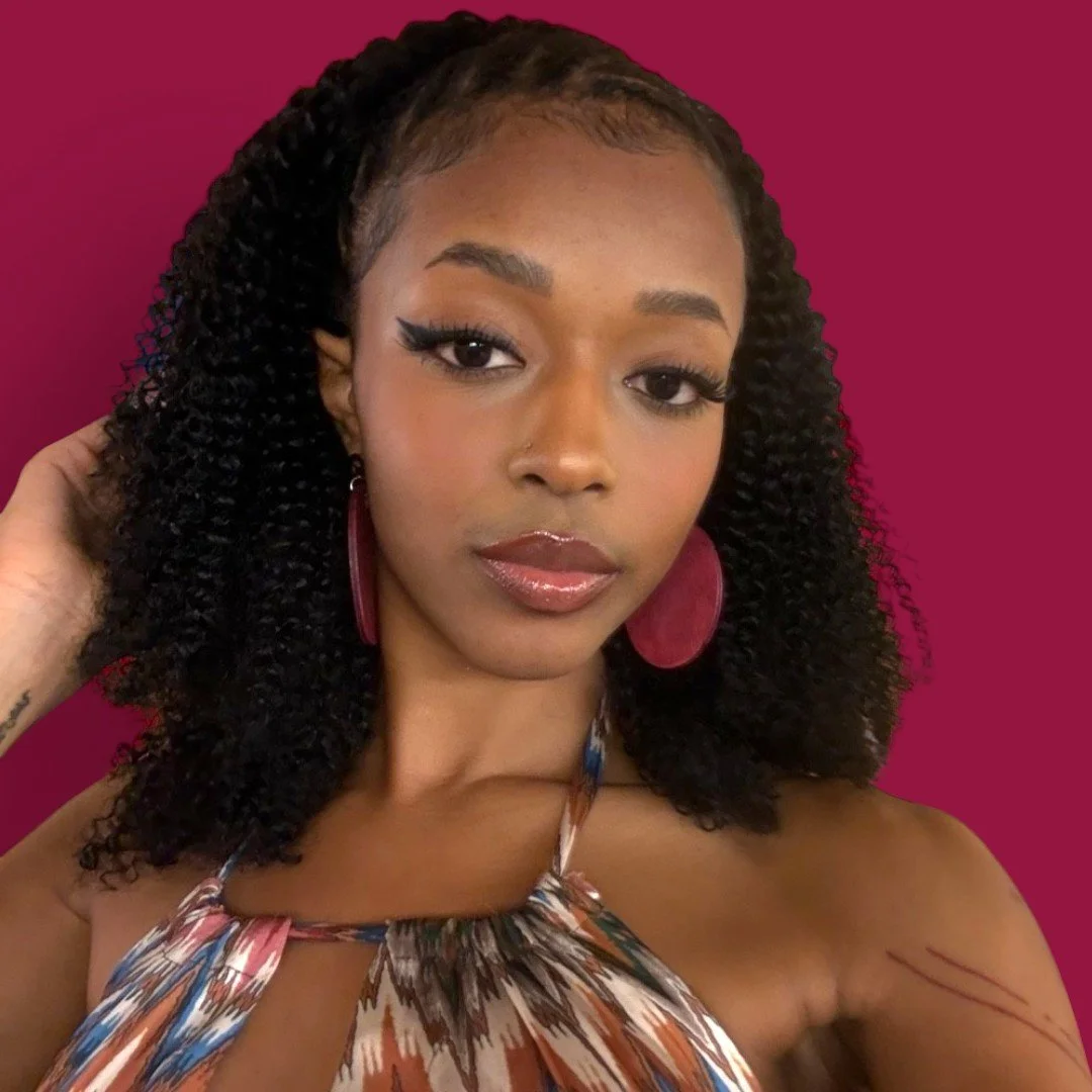 A young woman with curly hair and large earrings made by Color Cult and she is posing against a dark pink background.