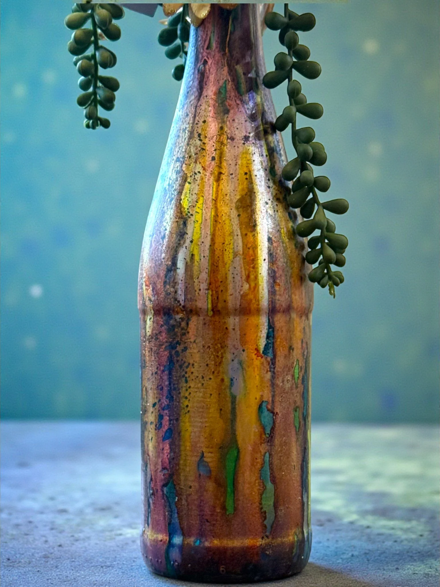 A colorful ceramic vase with dripping glaze in shades of yellow, orange, green, and blue, with green succulents hanging from the top.