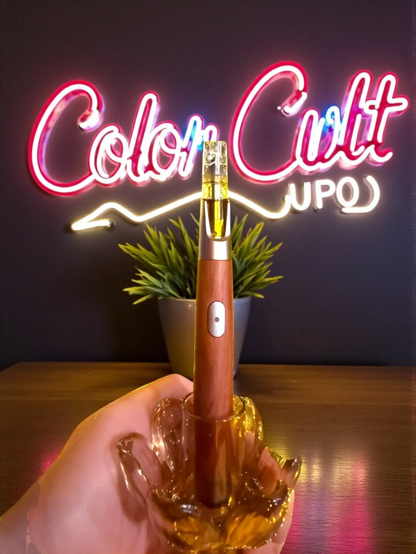 A hand holding a vaporizer pen with a vaporizer cartridge on top, in front of a neon sign that reads 'Color Cut' and a small potted plant in the background.