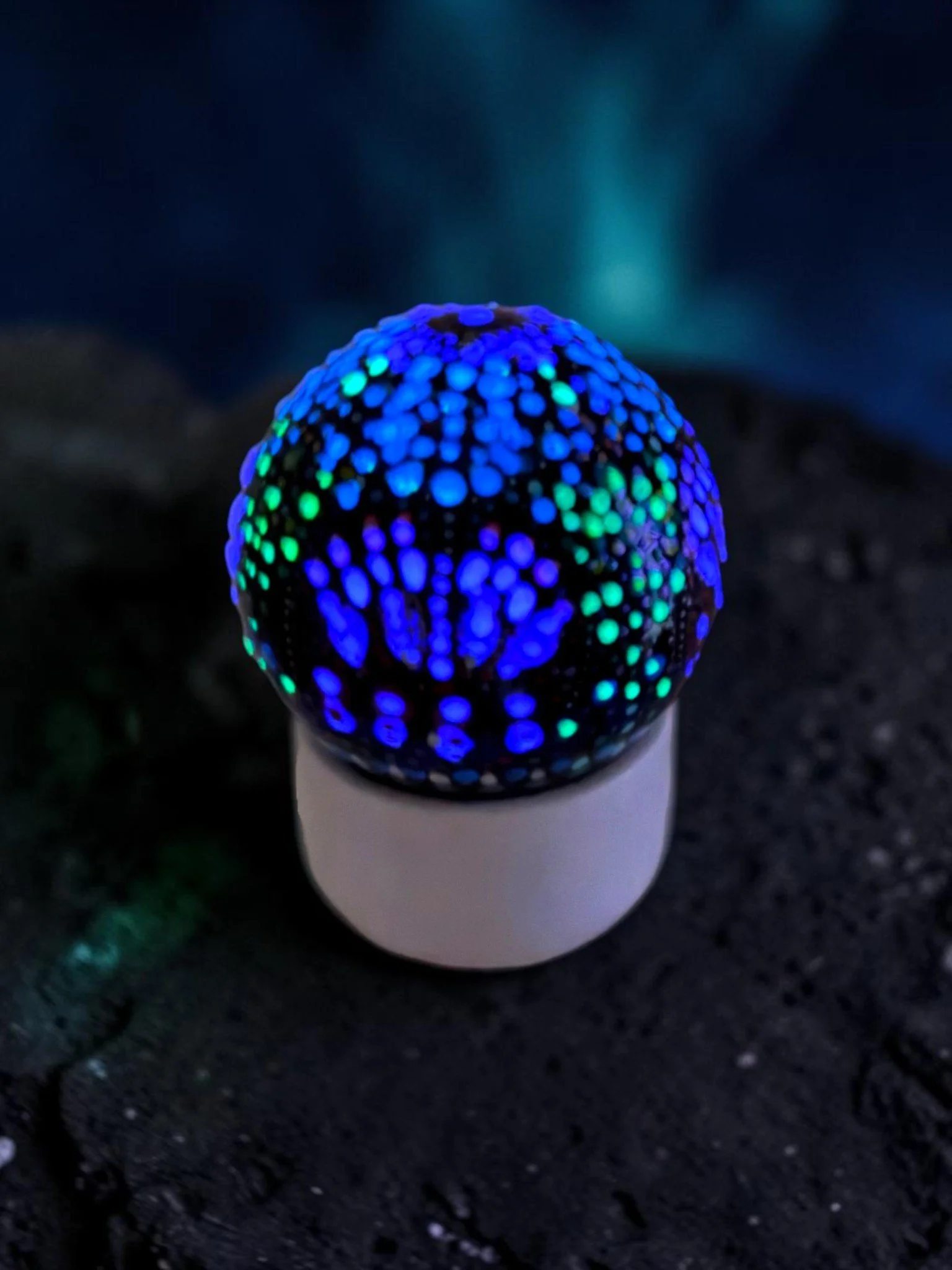 A glowing, colorful, ornamental glass globe with handpainted blue and green dots, affixed on top of a white pedestal base, photographed on a dark surface to show off the glow in the dark paint elements.