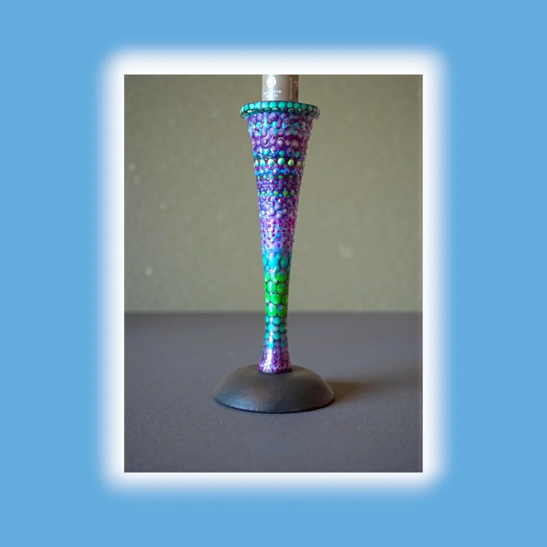Colorful, textured cone-shaped object with a base, decorated with beads or dots in shades of purple, blue, and green, standing on a gray surface.