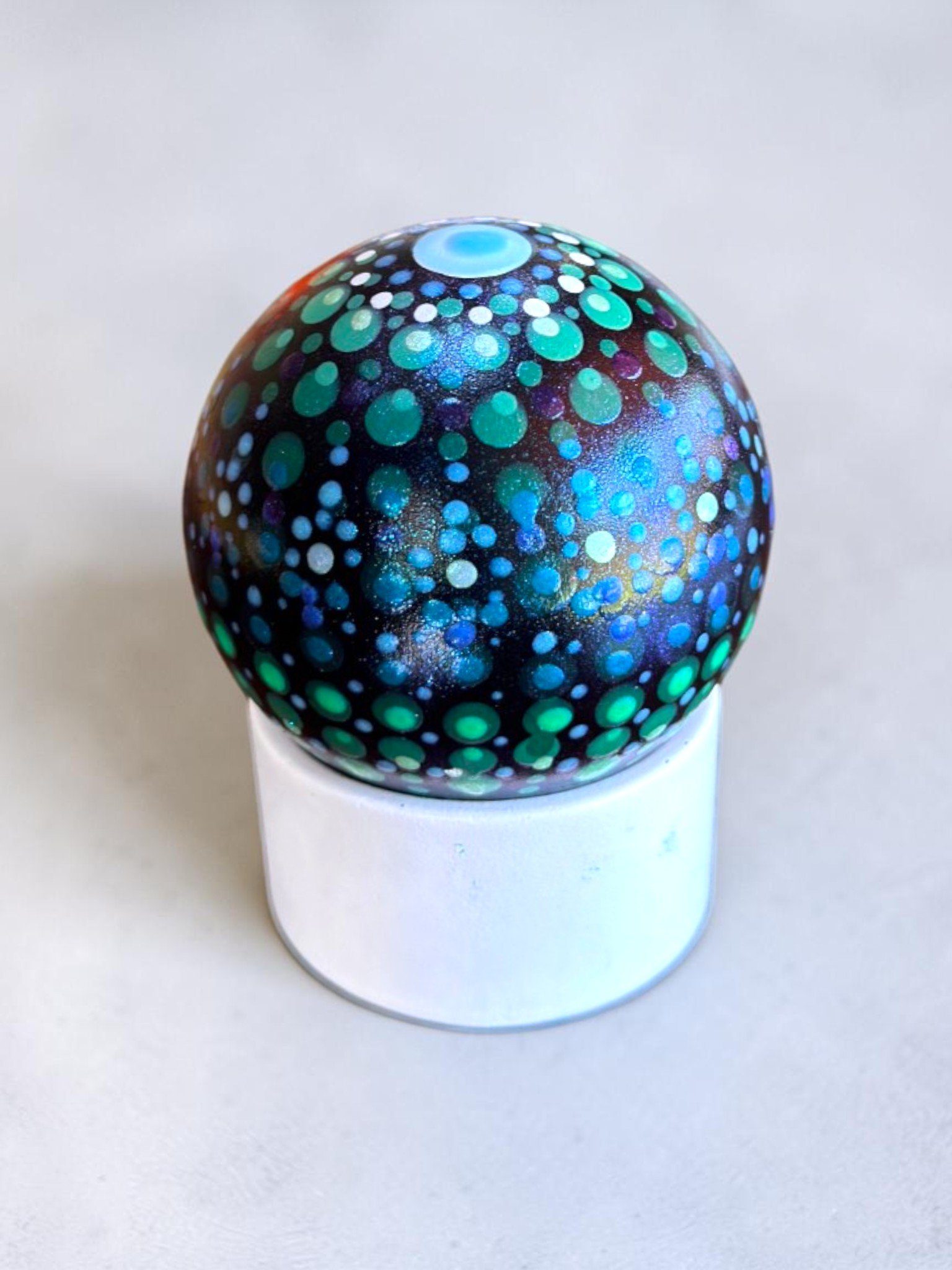 A decorative glass paperweight with a black background and colorful dot patterns in shades of blue, green, purple, and white, resting on a white stand.