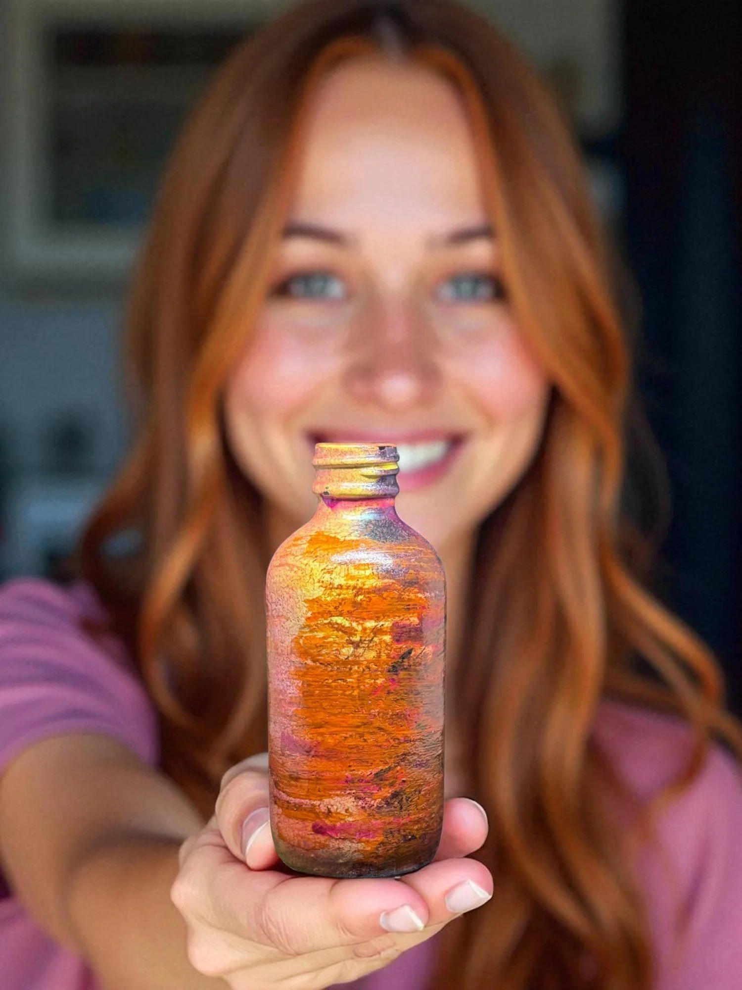 A woman with long red hair and a pink shirt smiling, holding a small jar with a painted sunset landscape on it, in front of her face.