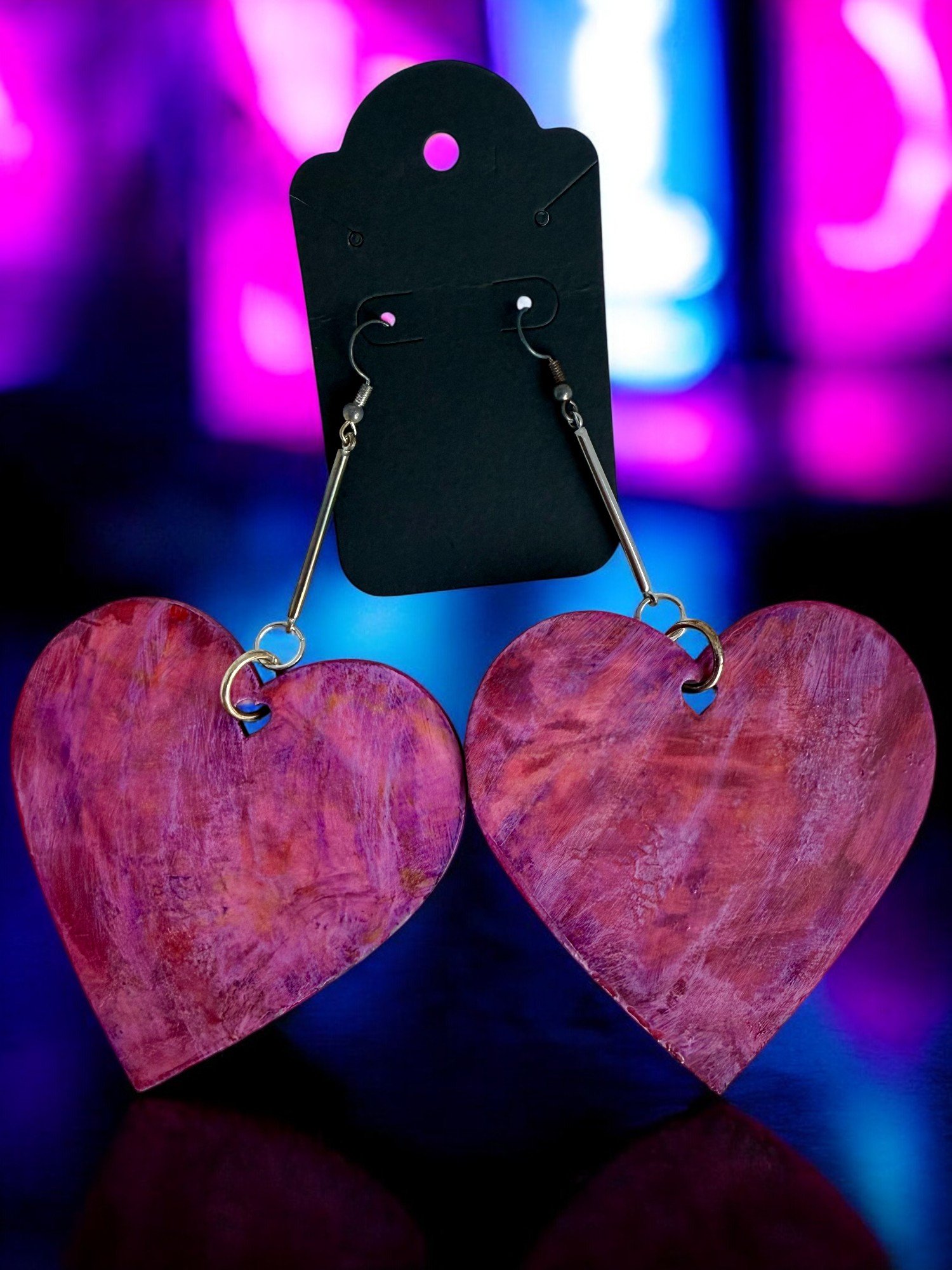 A pair of earrings with pink, textured heart-shaped pendants hanging from a black display card against a vibrant, colorful blurred background.