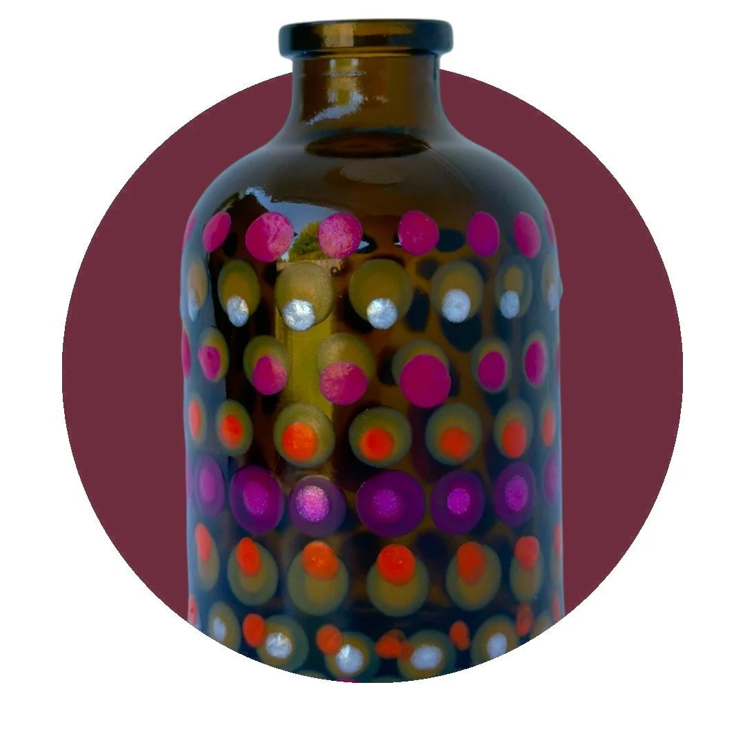 A multicolored glass bottle with a narrow neck, decorated with pink, silver, orange, and purple dots, on a maroon background.