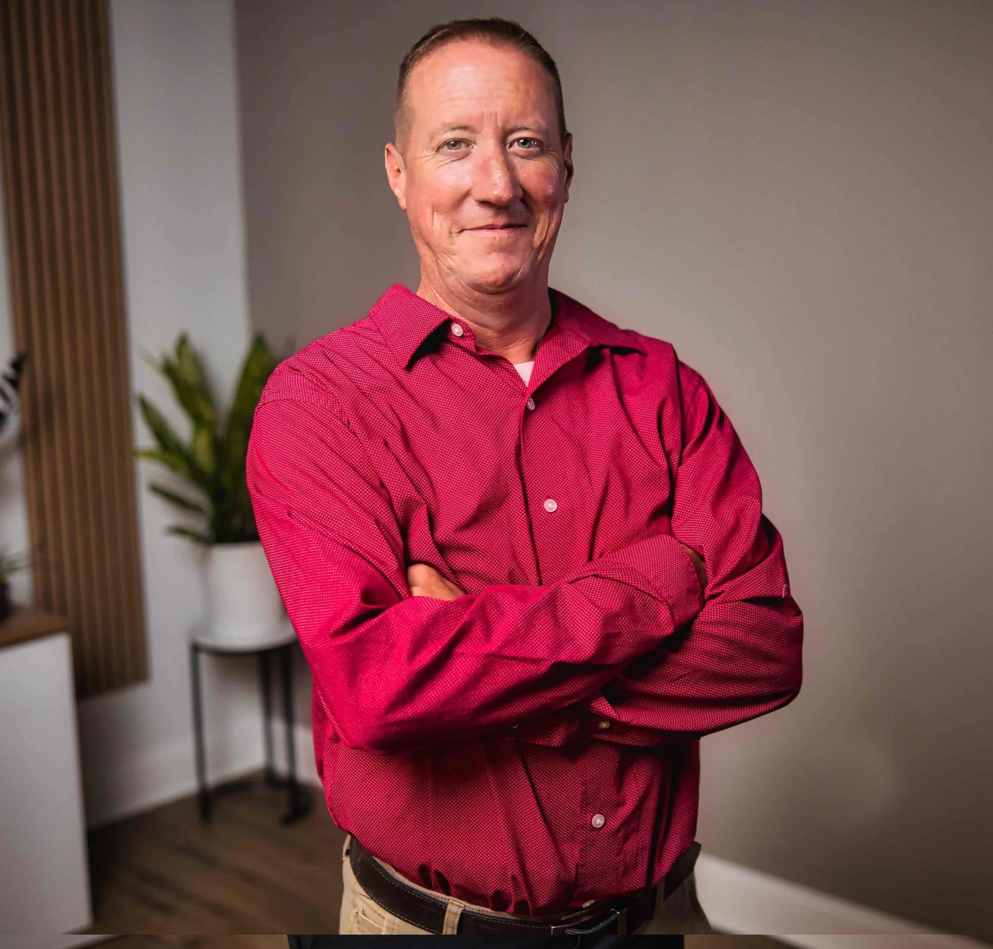 Man in red shirt crossing arms indoors, with plants and wood paneling in the background.