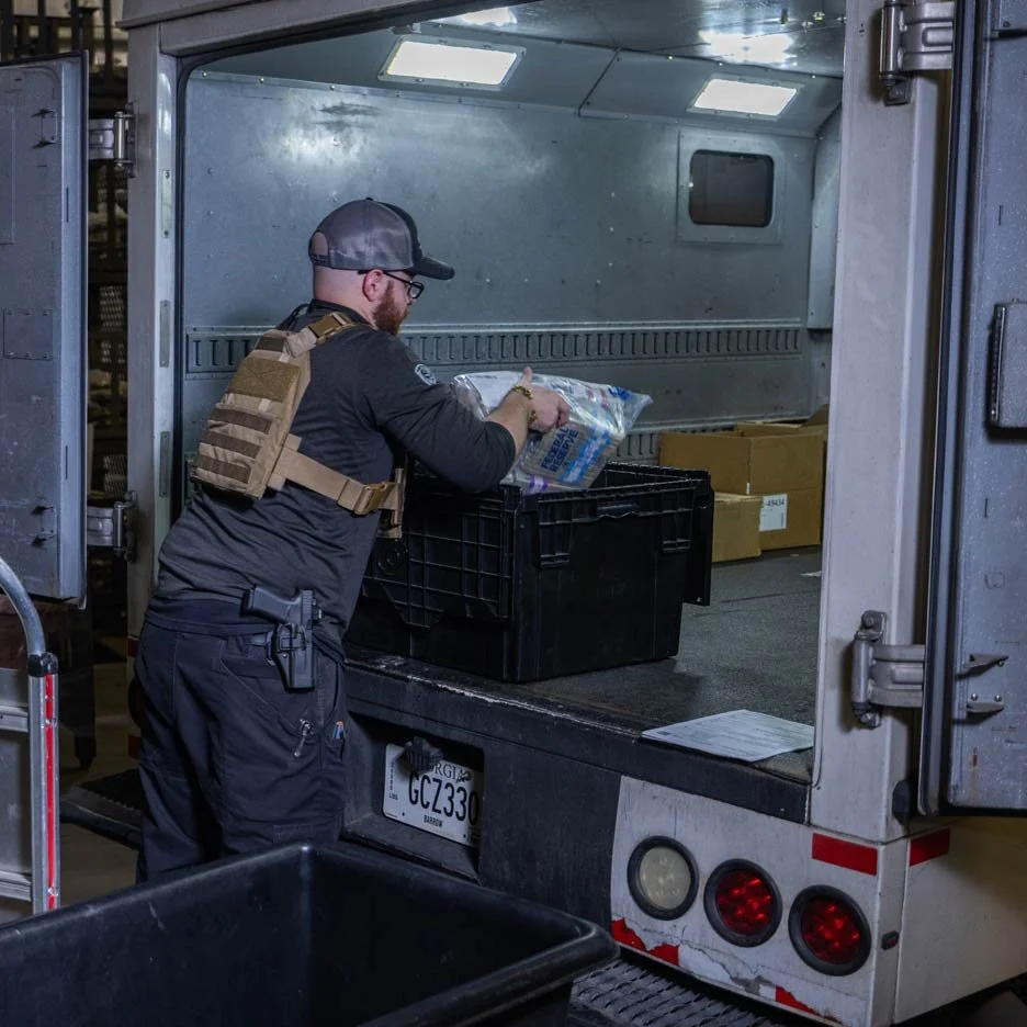 Man loading a crate into the back of a truck, wearing tactical gear and a cap, with boxes in the vehicle.