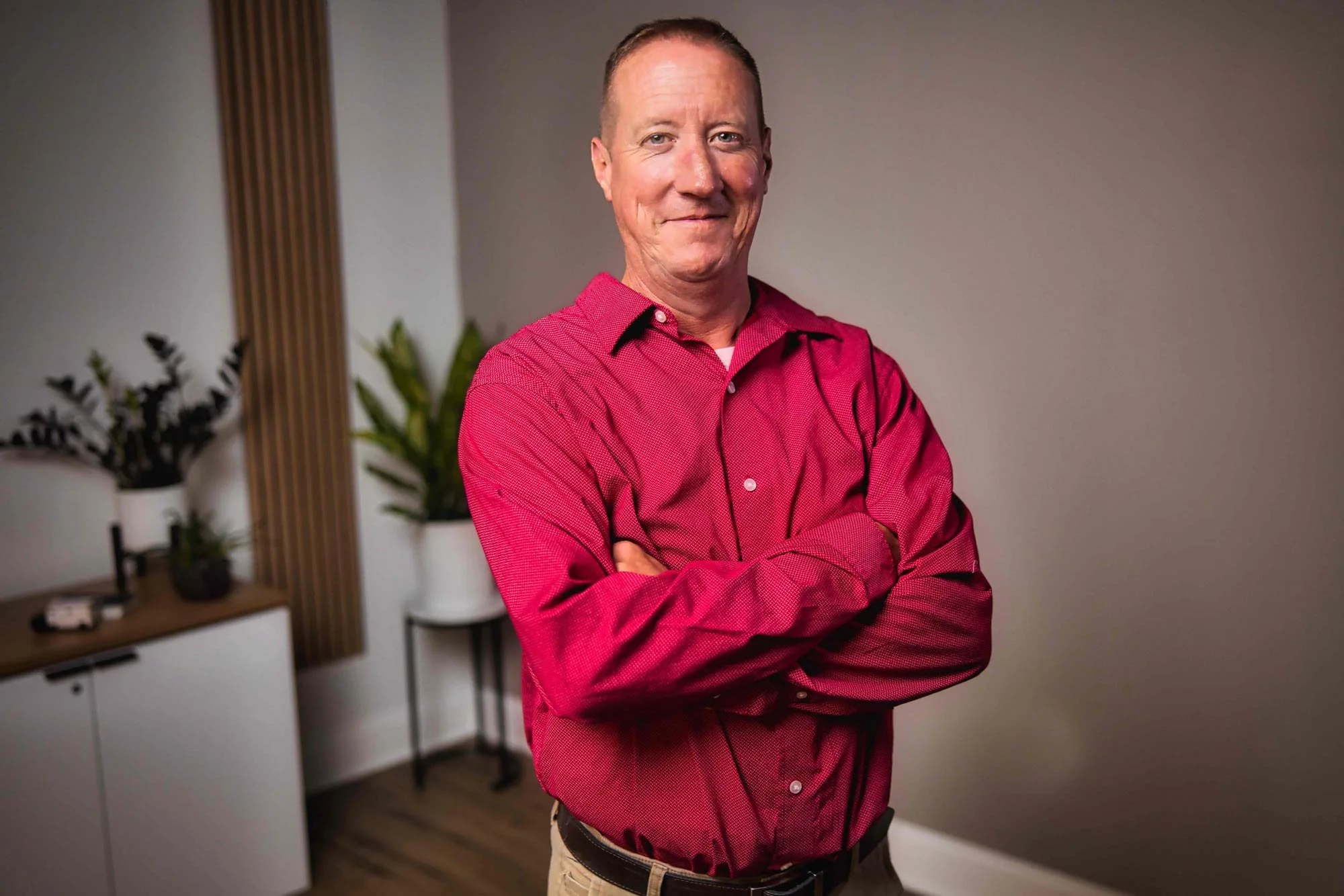 A man in a red shirt standing with arms crossed, smiling in a room with plants.