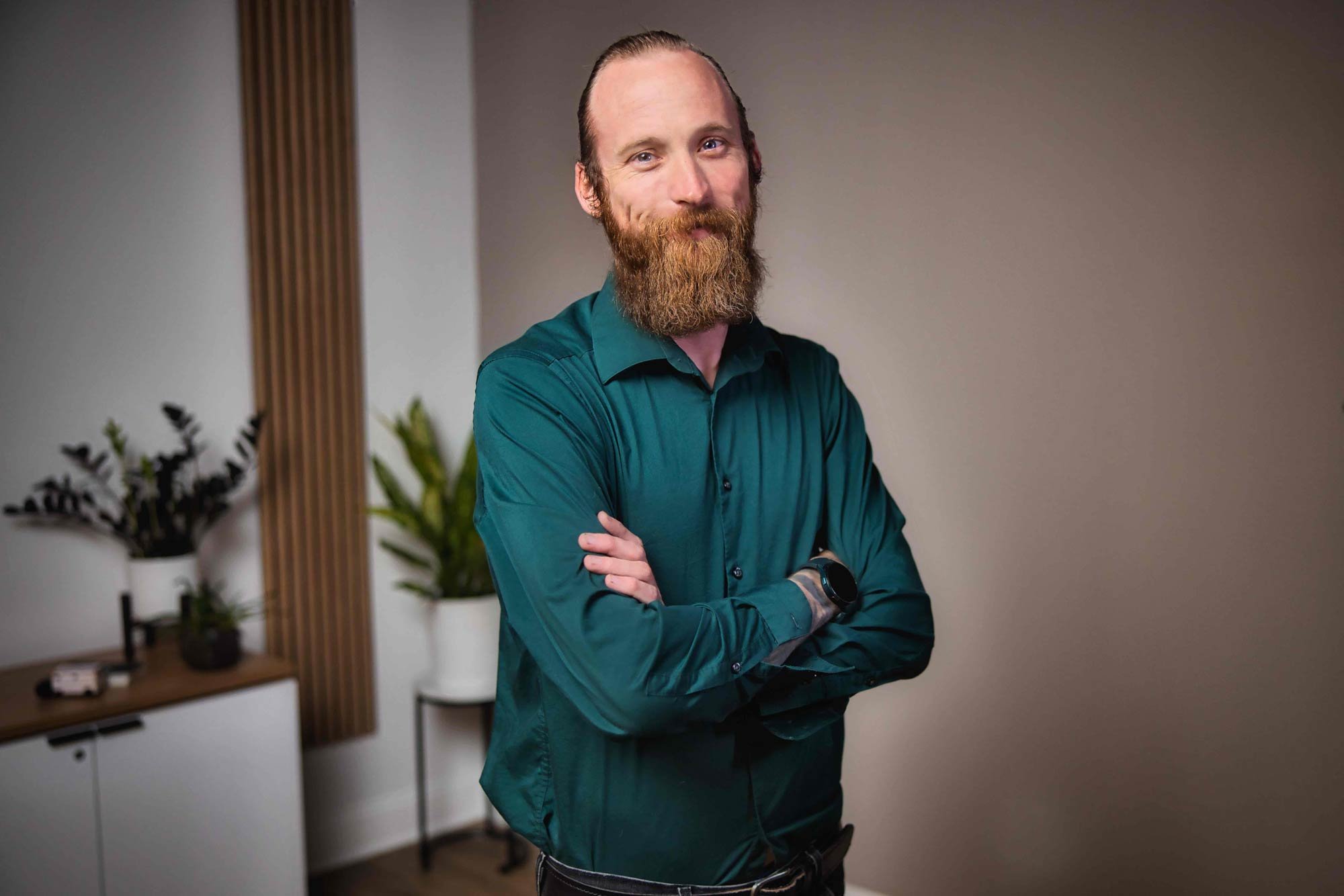 Person with a beard wearing a teal shirt, standing with crossed arms, indoor setting with plants in the background.