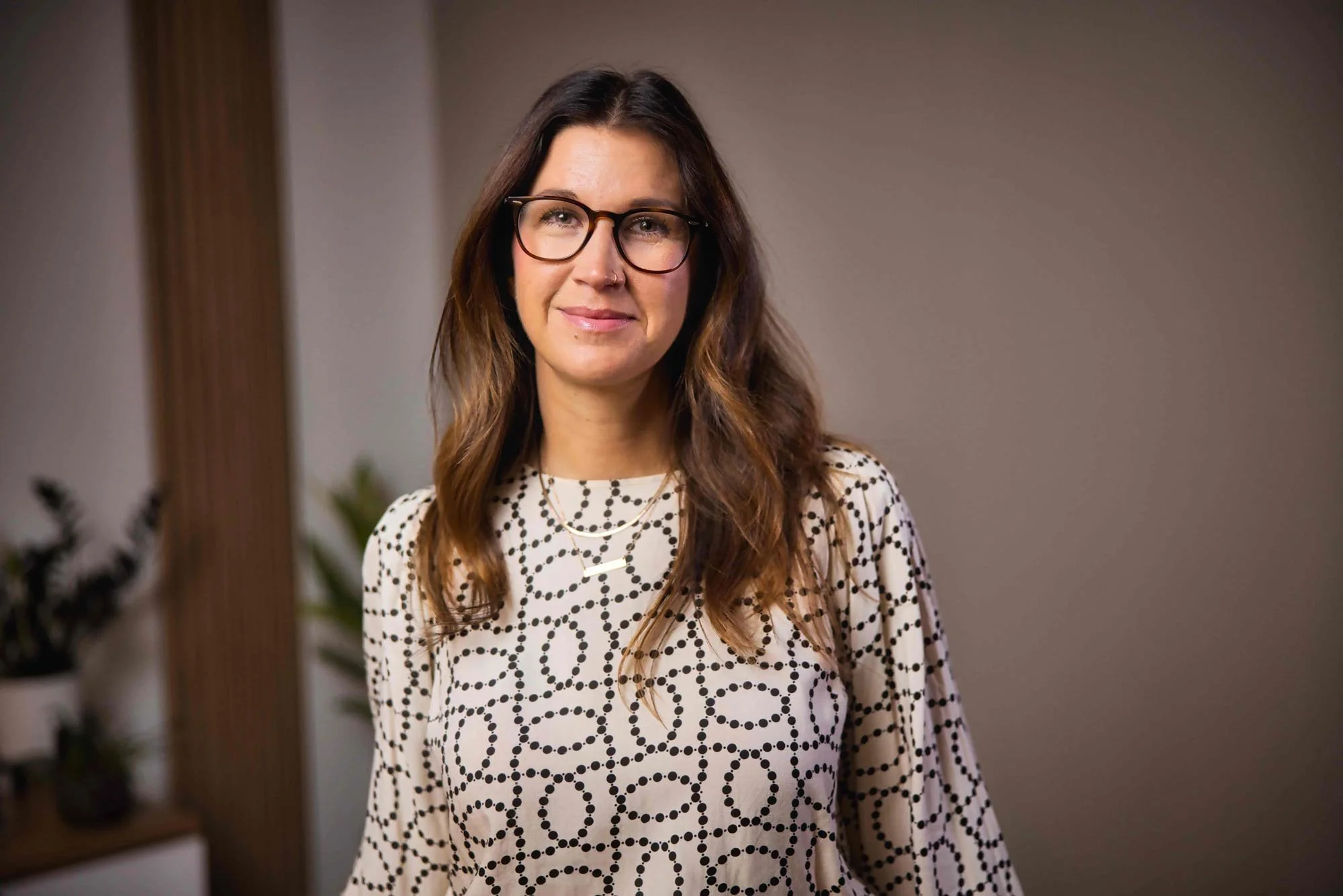 Woman with glasses in patterned blouse and long hair smiling indoors.