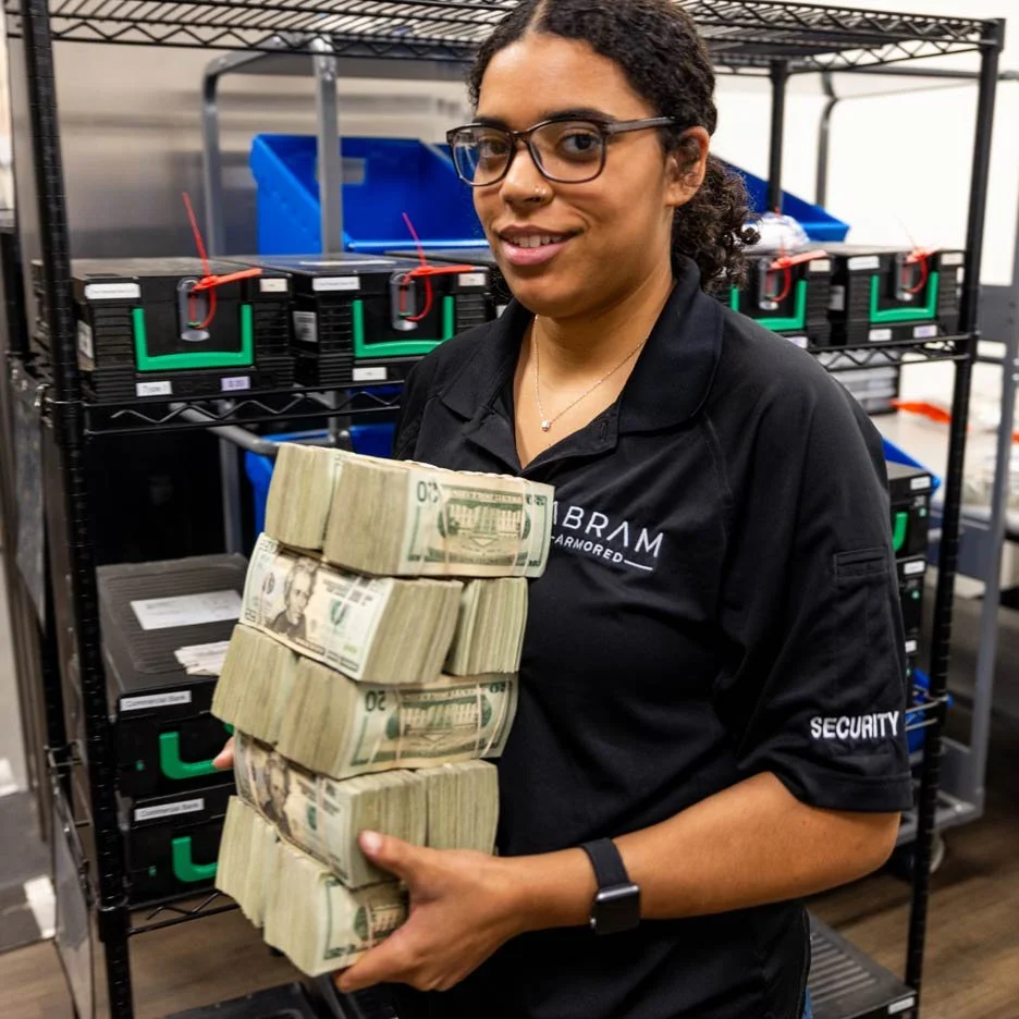 Person wearing a black security uniform holding stacks of U.S. dollar bills, standing in front of metal shelves with locked cash boxes.