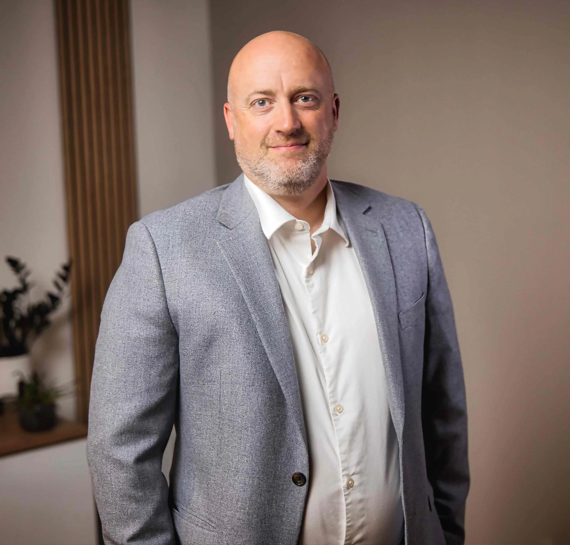 Man wearing a gray blazer and white shirt, standing indoors with plants in the background.