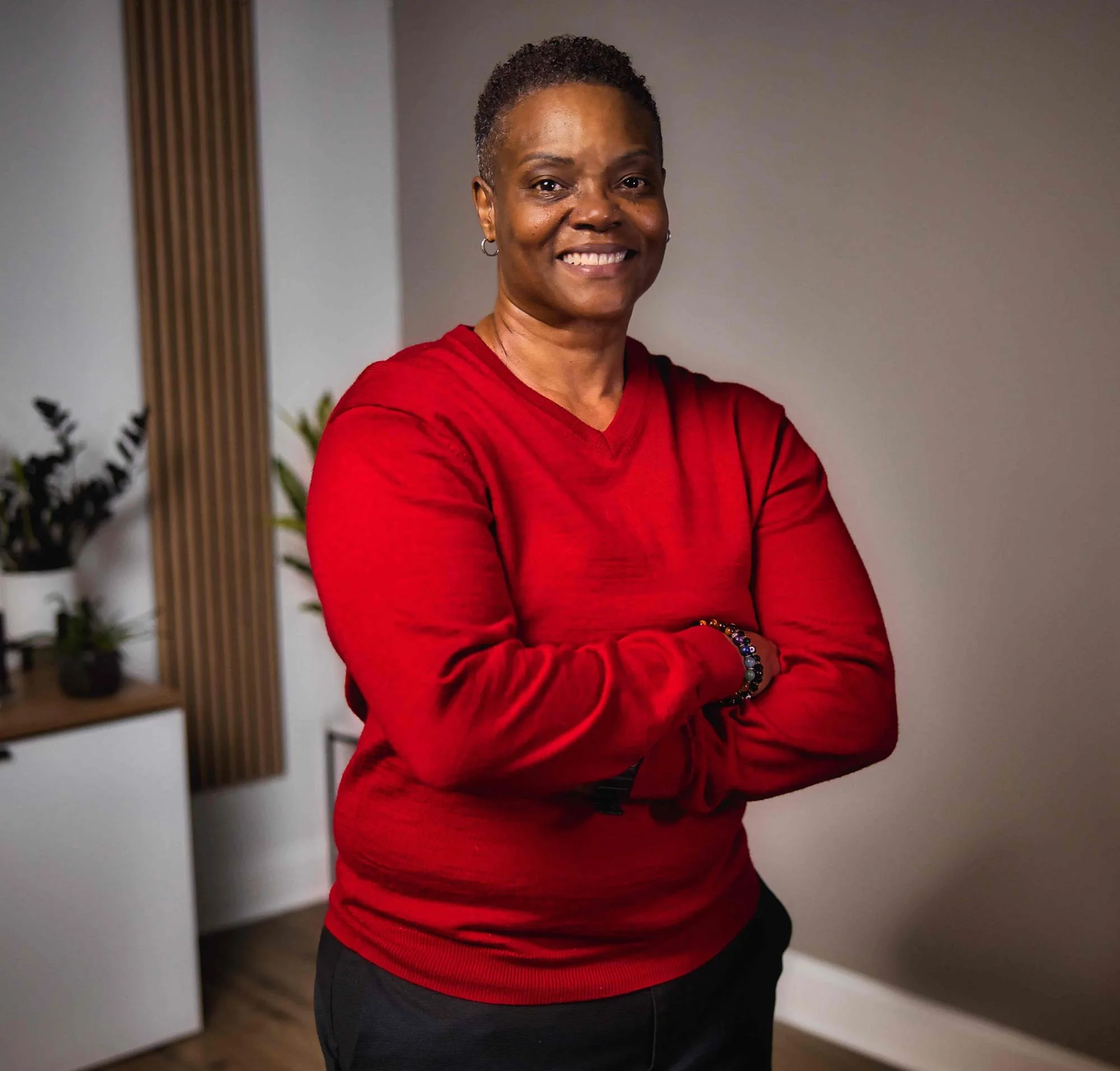 Smiling person in a red sweater standing with arms crossed in a well-lit room with plants and a cabinet in the background.