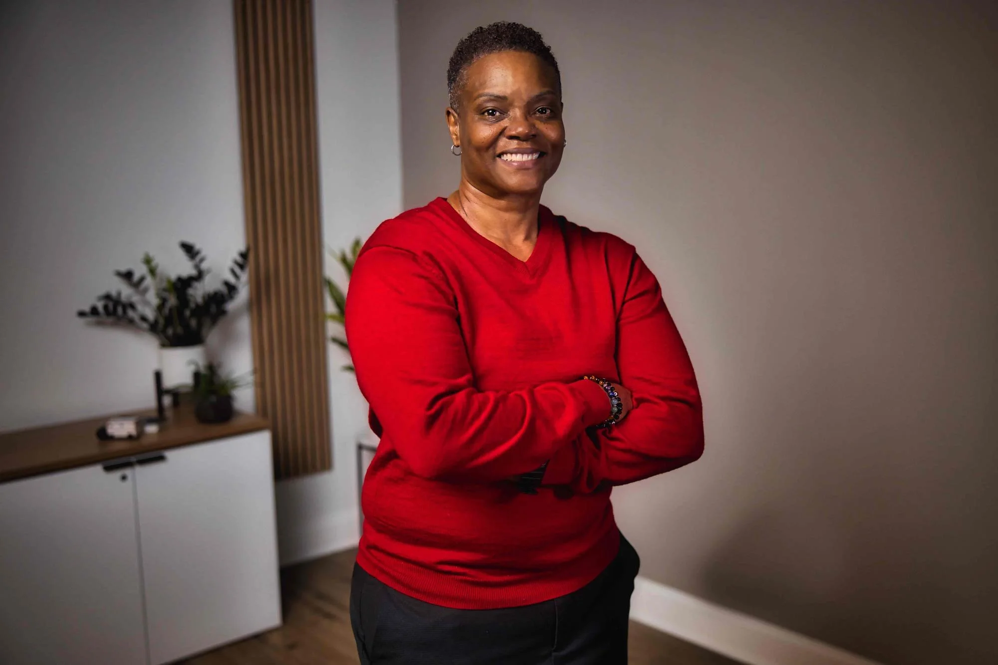 Person in red sweater smiling, crossed arms, indoors, plants and cabinet in background.