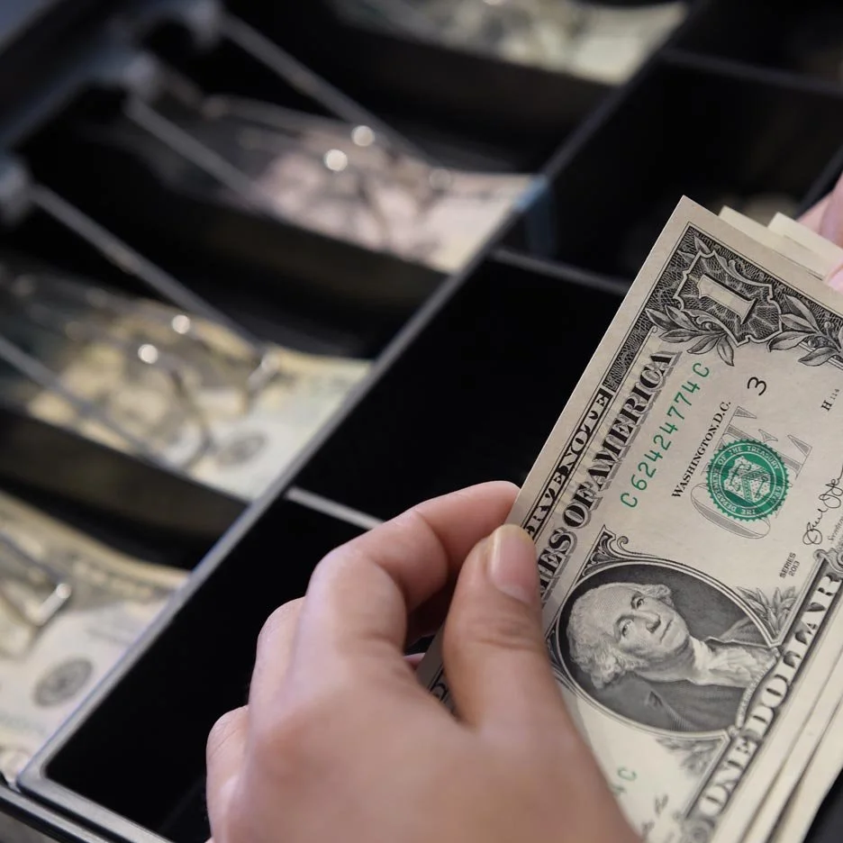 A person holding stacks of twenty-dollar bills in a secure room with shelves containing locked boxes and blue containers.