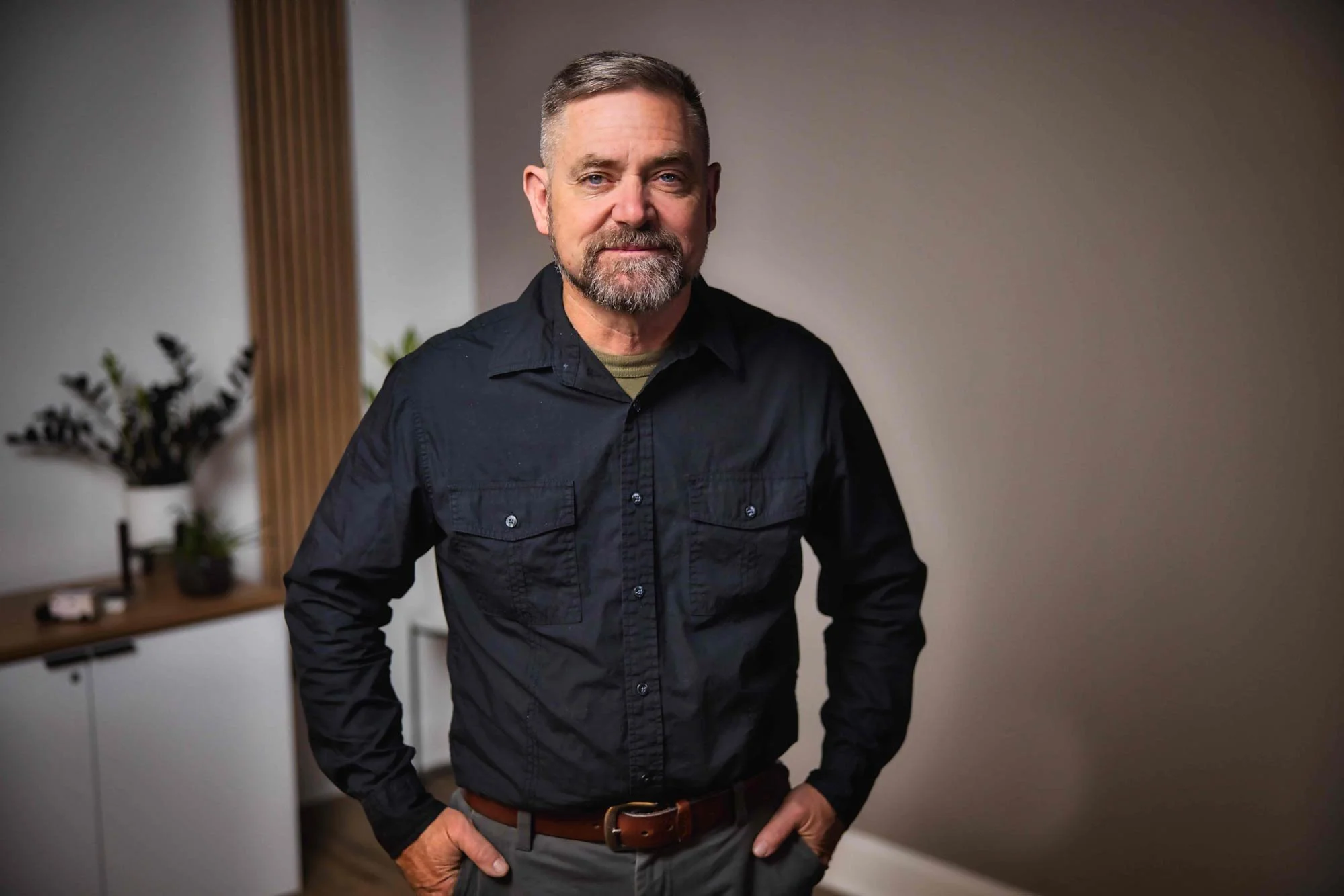 Adult male wearing a black shirt, standing indoors with a neutral expression. Background includes plants and a white cabinet.