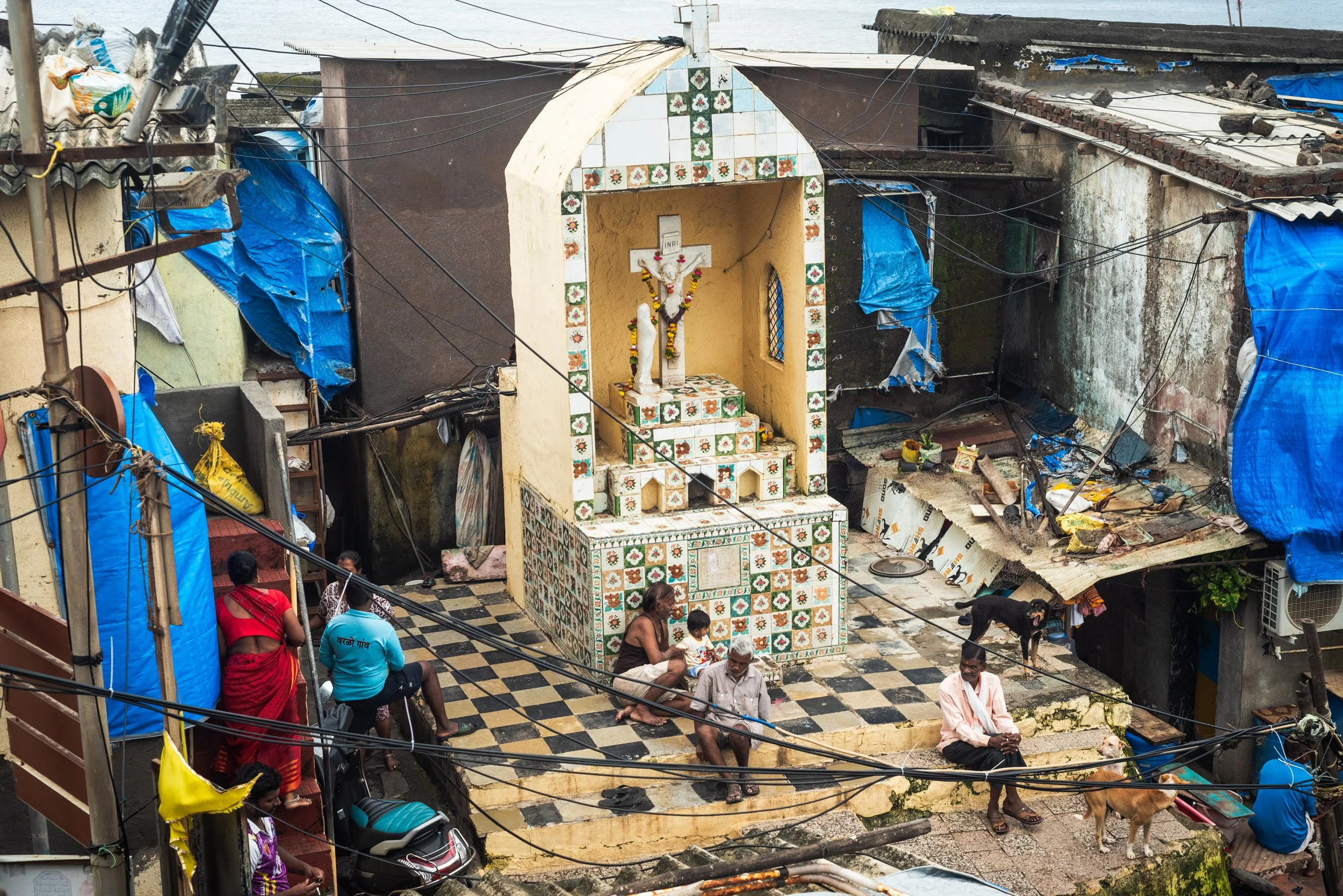 A small outdoor shrine with a tiled altar featuring a crucifix and religious statues, surrounded by worn buildings and people sitting and standing around, with some sitting on the steps and others near dogs, in an urban setting.