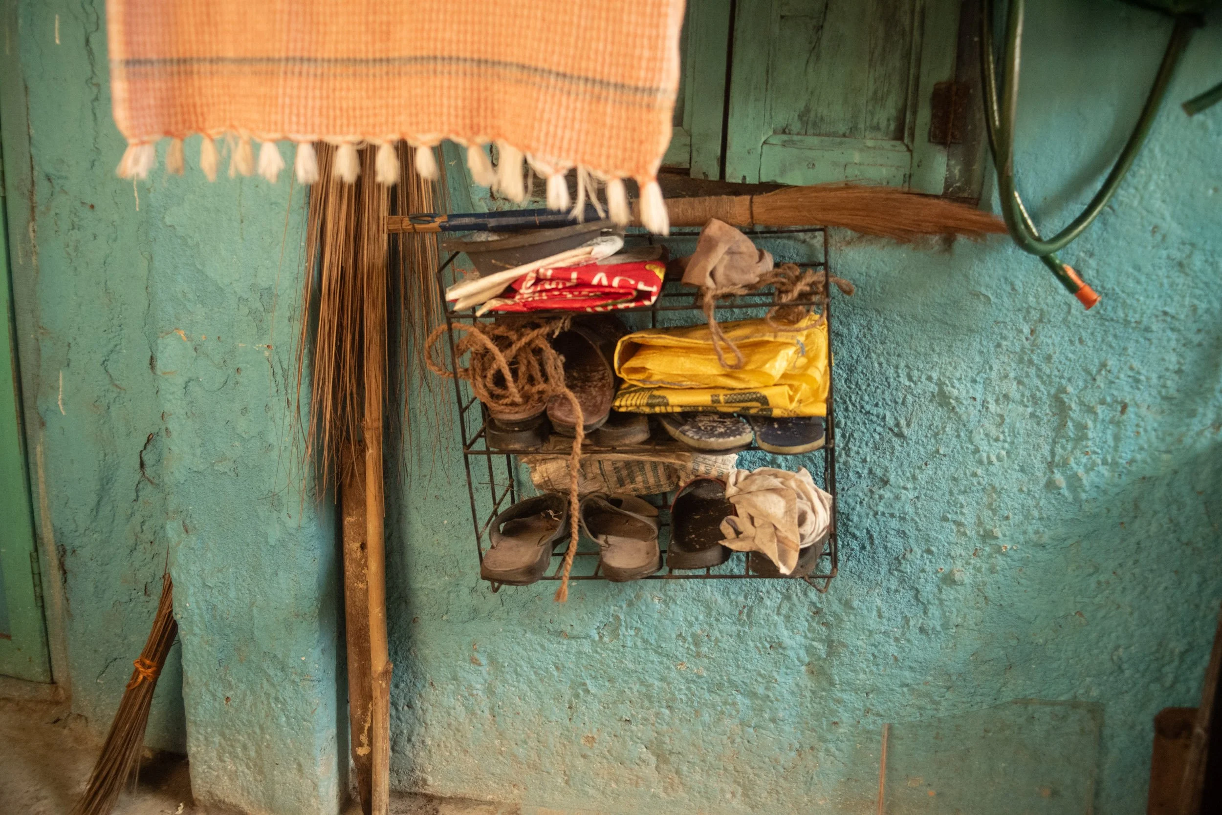 A wall-mounted metal shoe rack holding several shoes and sandals of various sizes and styles, with a yellow plastic bag and some cloth items on top, against a textured teal wall.