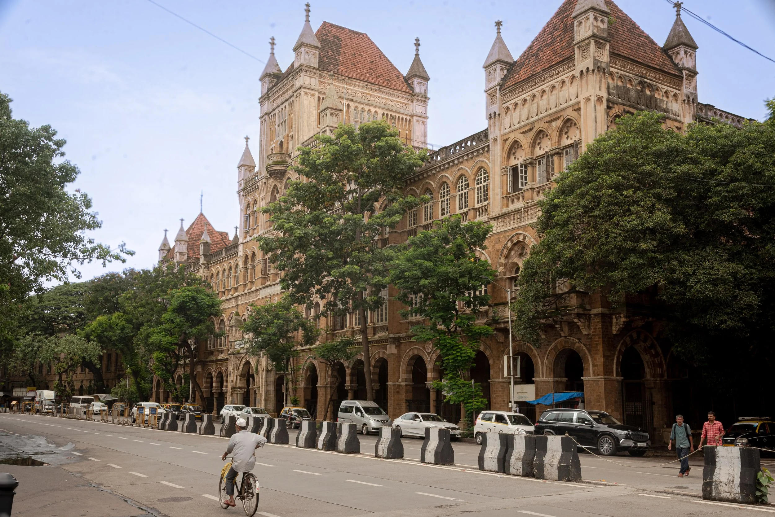 A historic castle-like building with multiple towers and arched windows, partially obscured by large green trees, with a street in front where people are walking and a cyclist is riding, cars parked along the side.