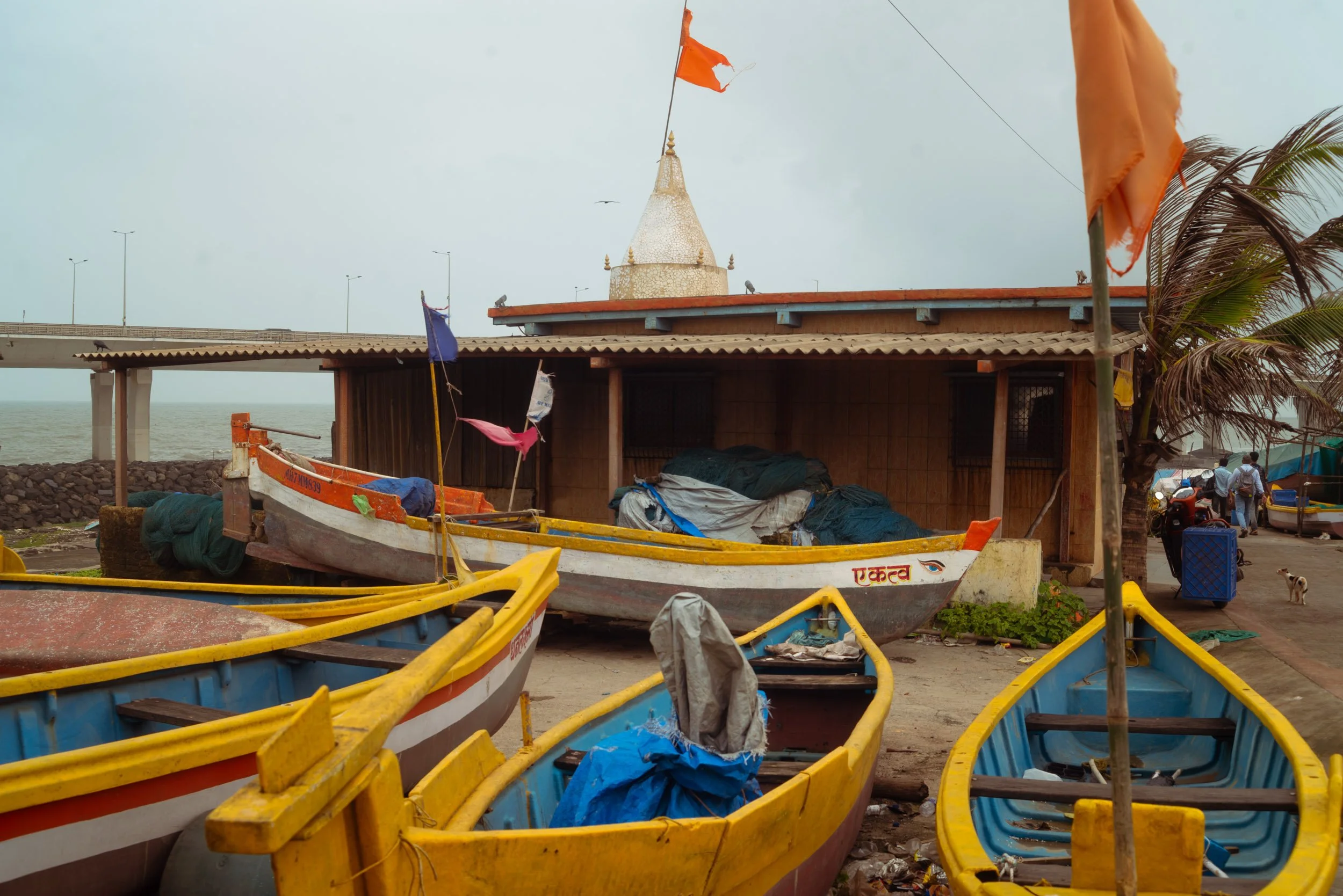 Colorful boats on the shore in front of a small building with flags and a temple-like structure on the roof, with a bridge and ocean in the background.