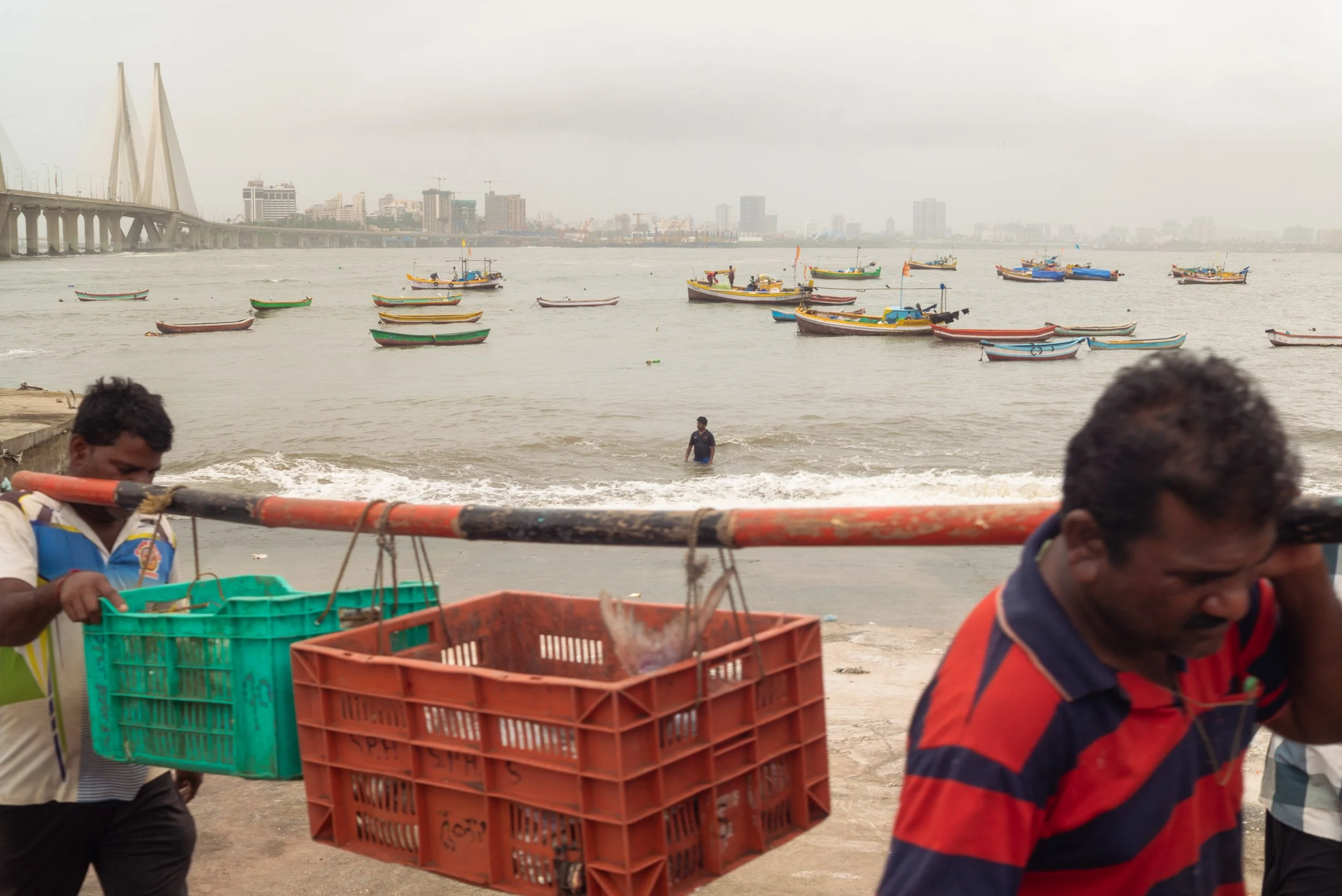 Mens at a fishing dock with boats on the water and city skyline in the background.