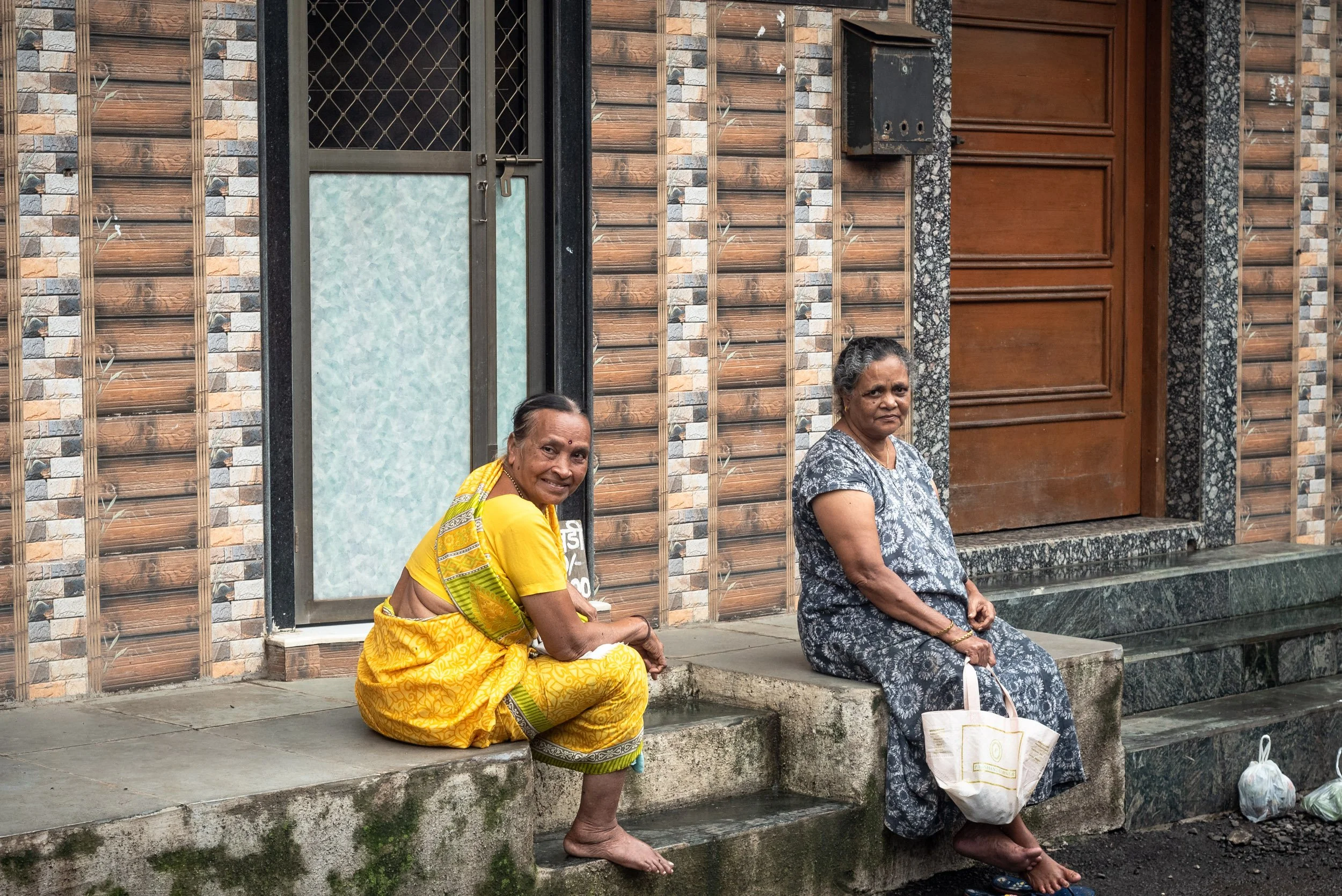 Two women sitting on a concrete step outside a building with a tiled wall. One woman wears a yellow sari and smiles, the other wears a grey patterned dress and looks serious.