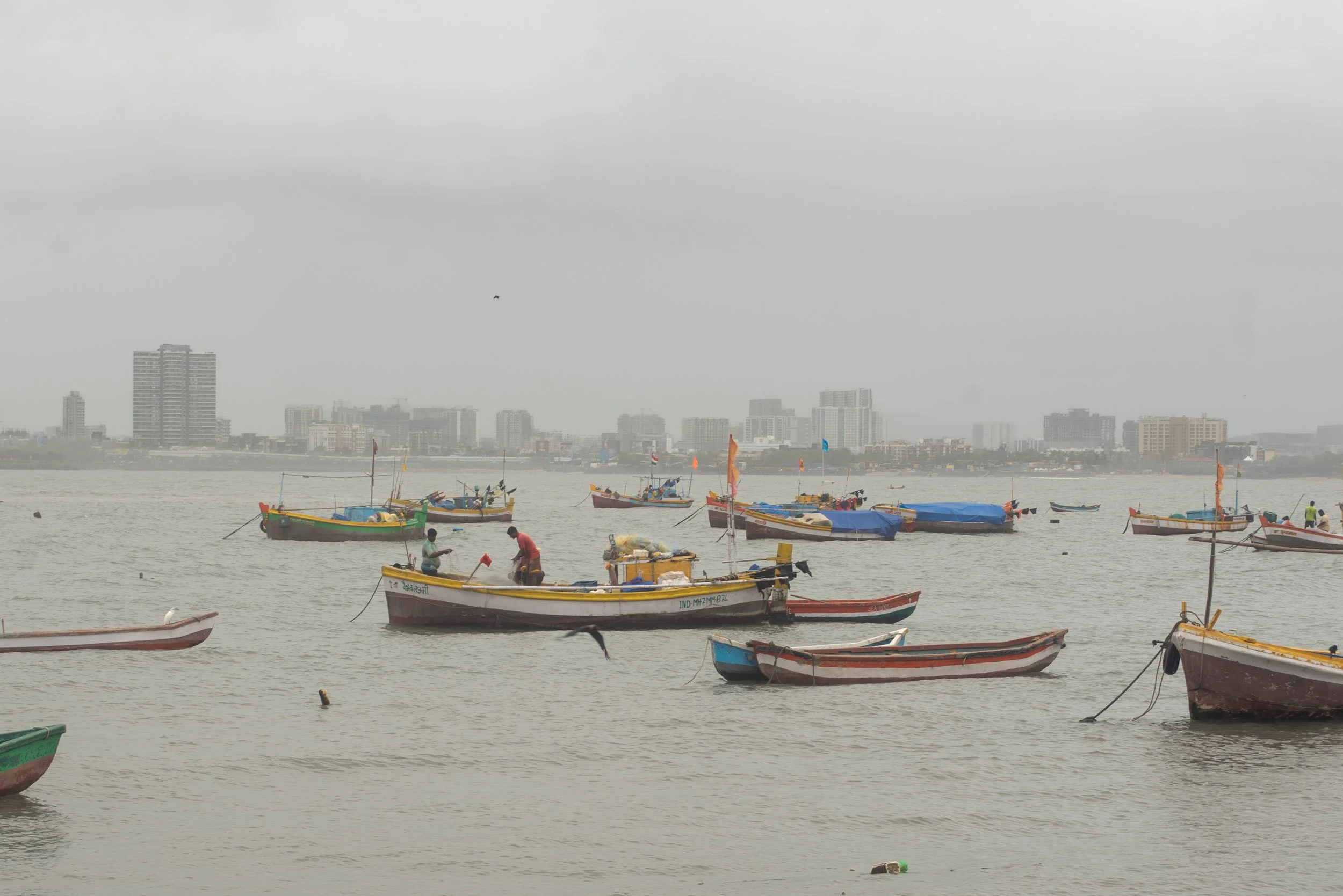 Multiple small boats floating on a calm water body with a cityscape of tall buildings and overcast sky in the background.