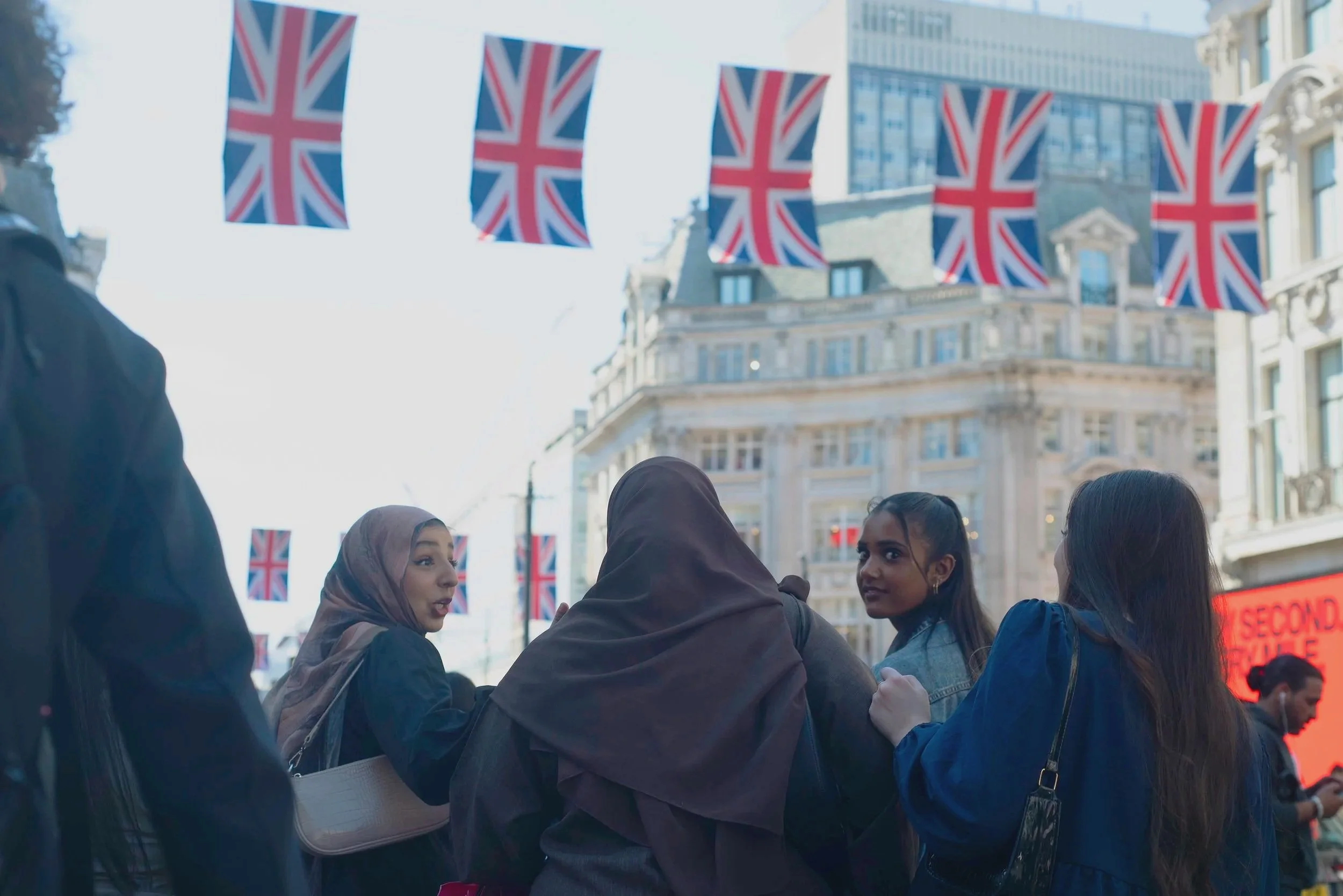 Group of diverse people gathered outdoors under Union Jack flags in a city street, with historic buildings and a digital billboard in the background.