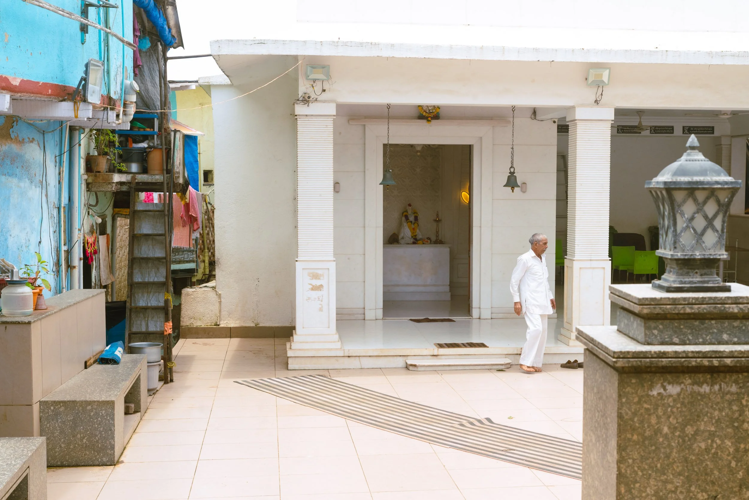 An elderly man dressed in white is walking near a small temple or shrine with a deity statue inside, outside the temple courtyard. The courtyard has tiled flooring, a welcome mat, and a bench with household items on the left. To the left side, there 