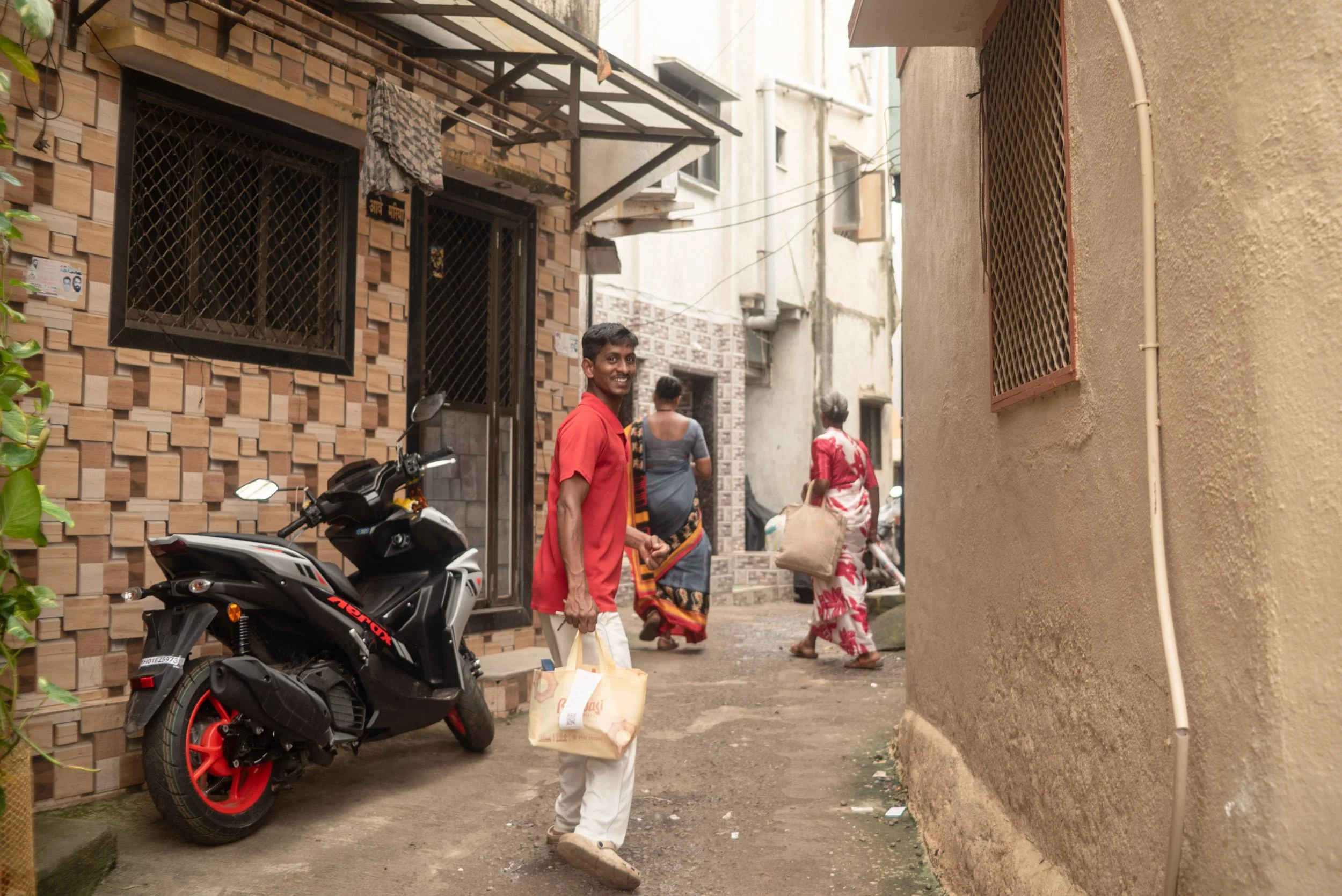 A man in a red shirt smiling while holding a paper bag walks down a narrow alleyway between buildings in an urban area. Two women, one in a gray sari and the other in a red and white sari, walk ahead carrying bags. A black and red motorcycle is parke
