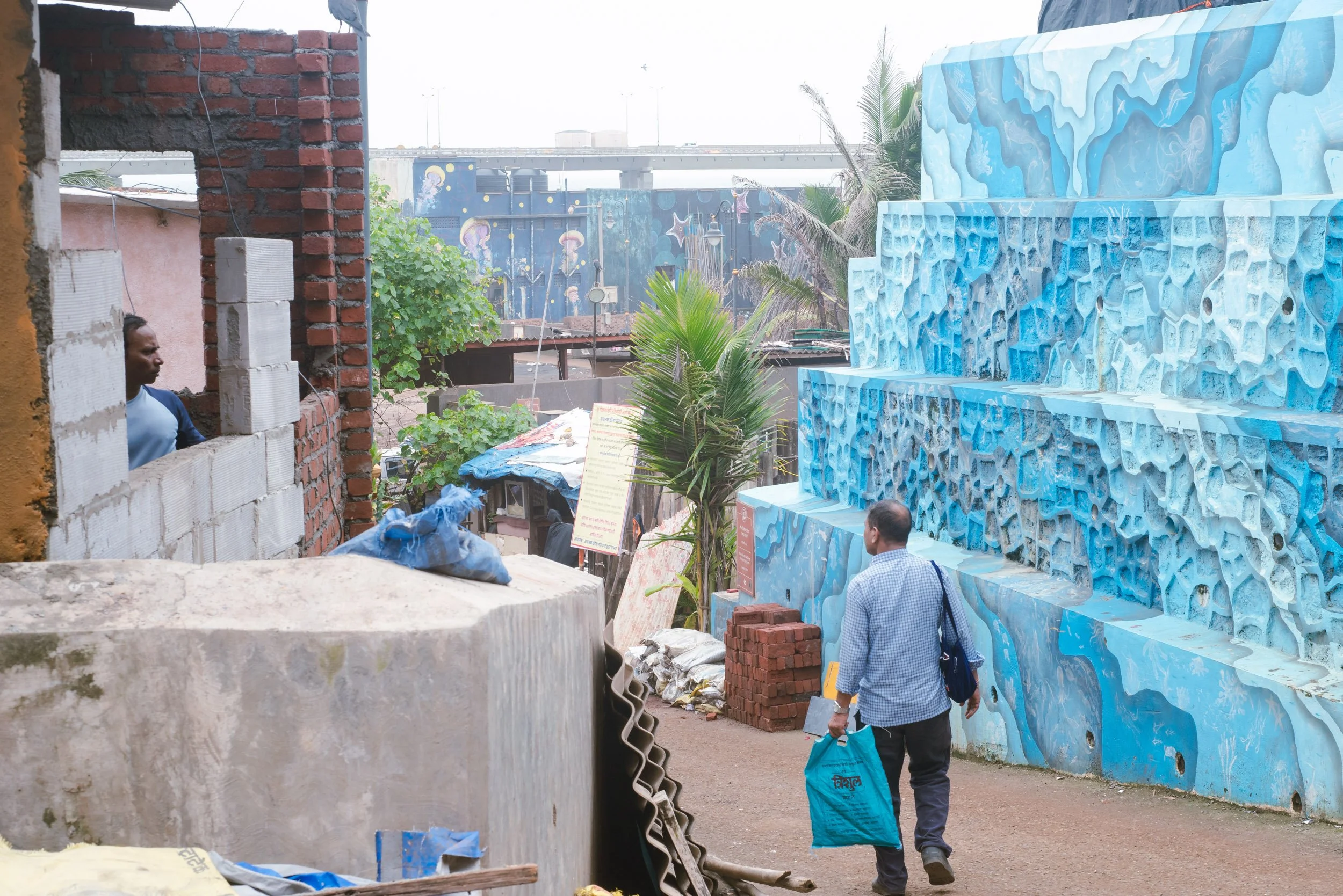 A man walking on a dirt path carrying a blue bag in an alley, with a wall of blue sculptures on his right and a partially constructed brick wall on his left, in an urban area.