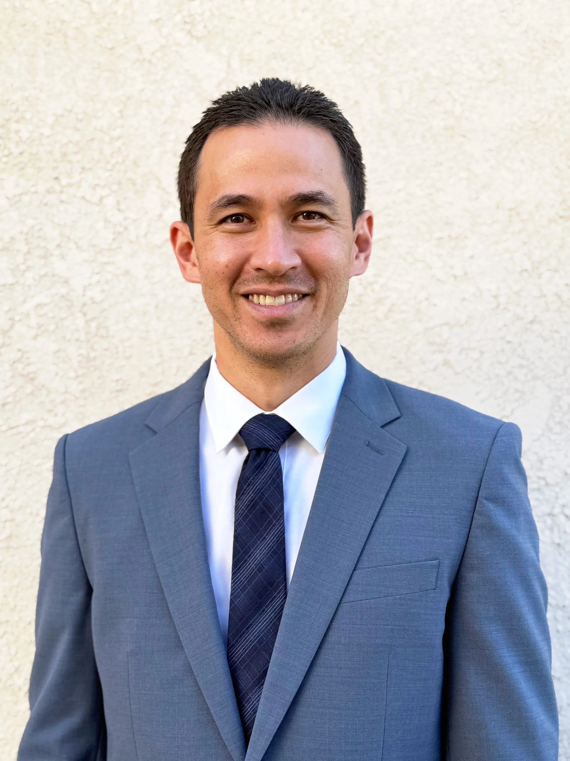 A man in a gray suit, white shirt, and navy tie smiling in front of a cream-colored wall.