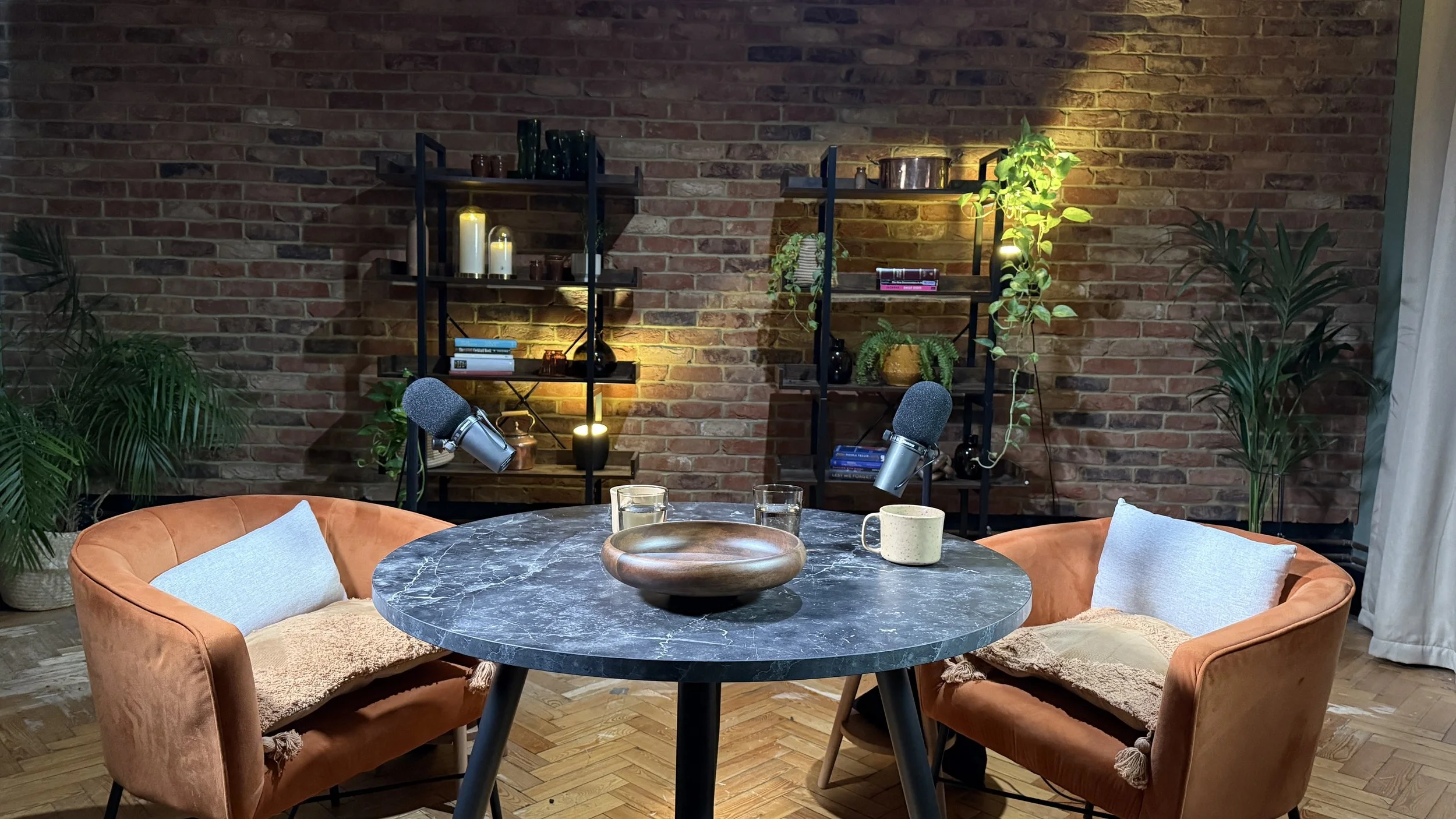 Cozy podcast recording setup with two orange velvet armchairs with pillows, a black marble round table with a wooden bowl, cups, and glasses, two microphones positioned towards the chairs, and a brick wall background with shelving holding candles, bo