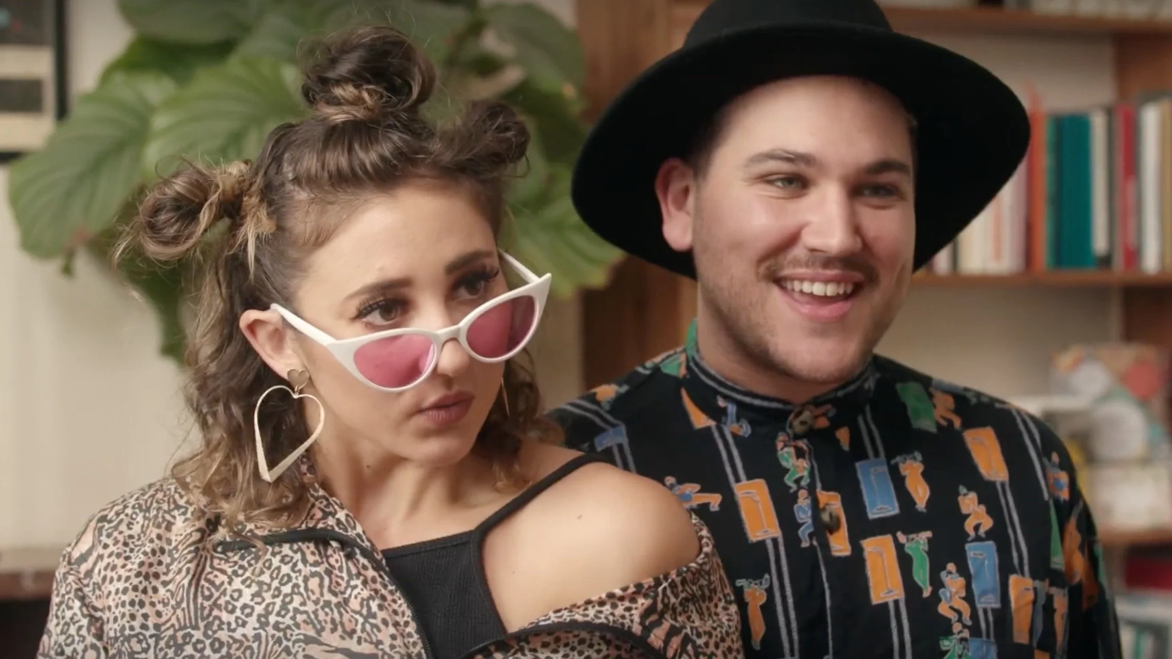 Two young adults, a woman and a man, standing indoors with bookshelves and greenery in the background. The woman has curly hair styled in two buns, wears pink-tinted sunglasses, a leopard print top with an off-shoulder black shirt underneath, and lar