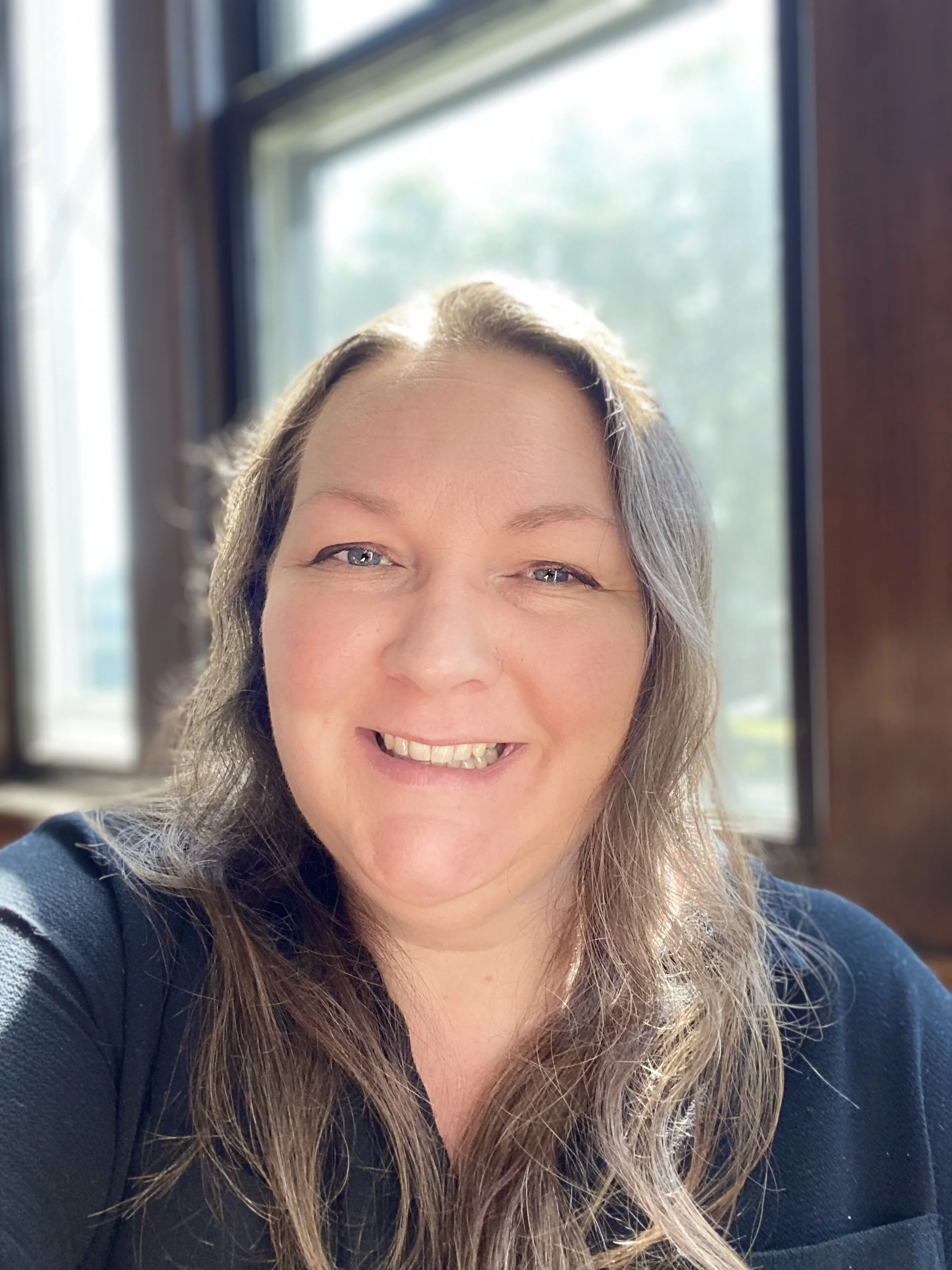 A smiling woman with long brown hair sitting near a window with sunlight streaming in.