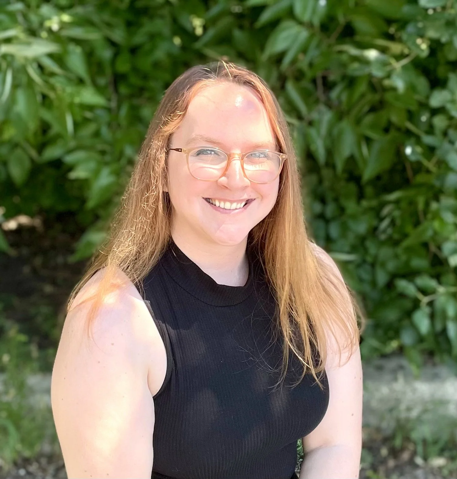 A woman with long red hair wearing glasses and a black sleeveless top, smiling outdoors in front of green foliage.