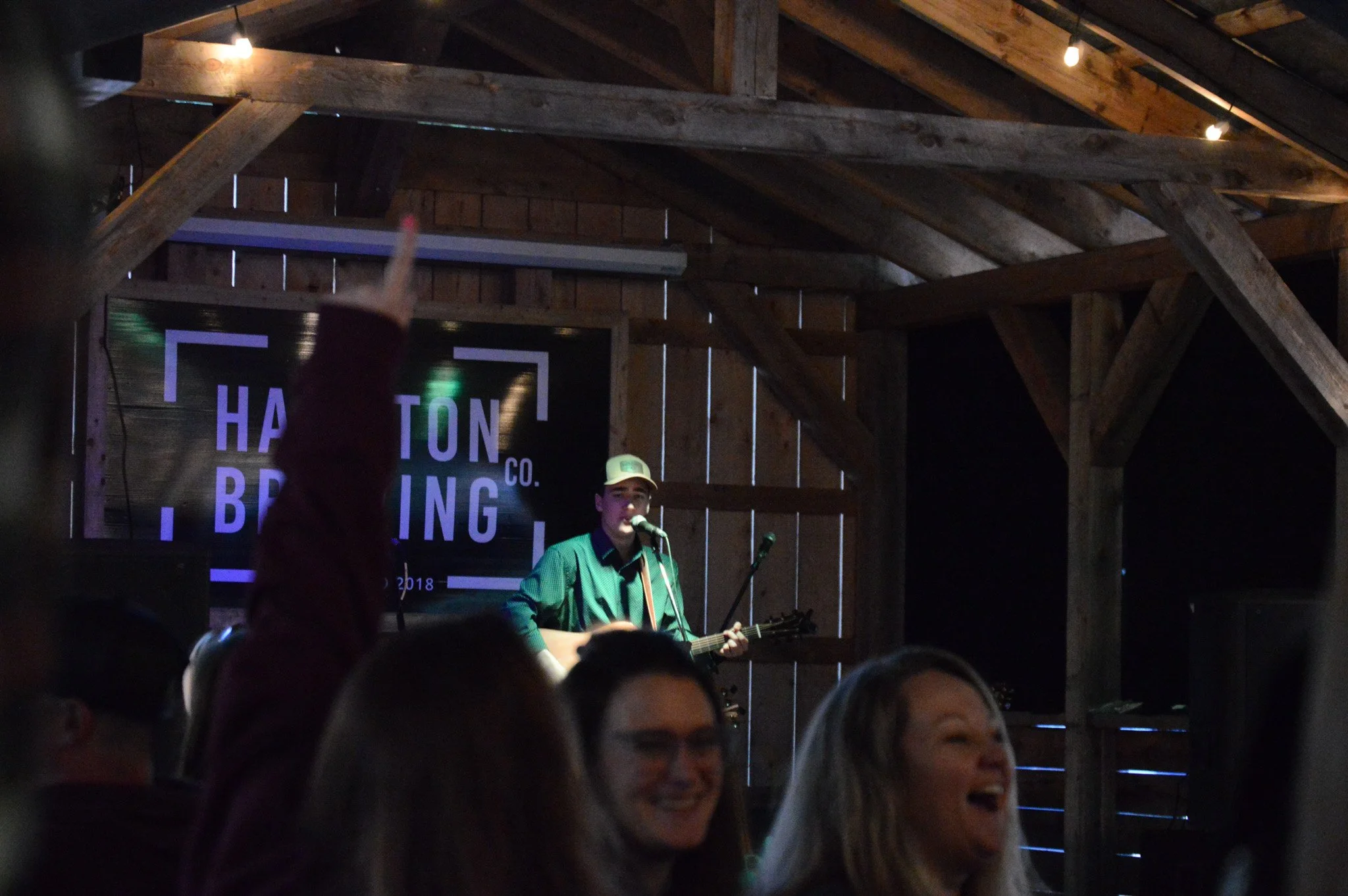 A man is performing on stage with a guitar and singing into a microphone in a rustic wooden venue, while audience members are enjoying the show.