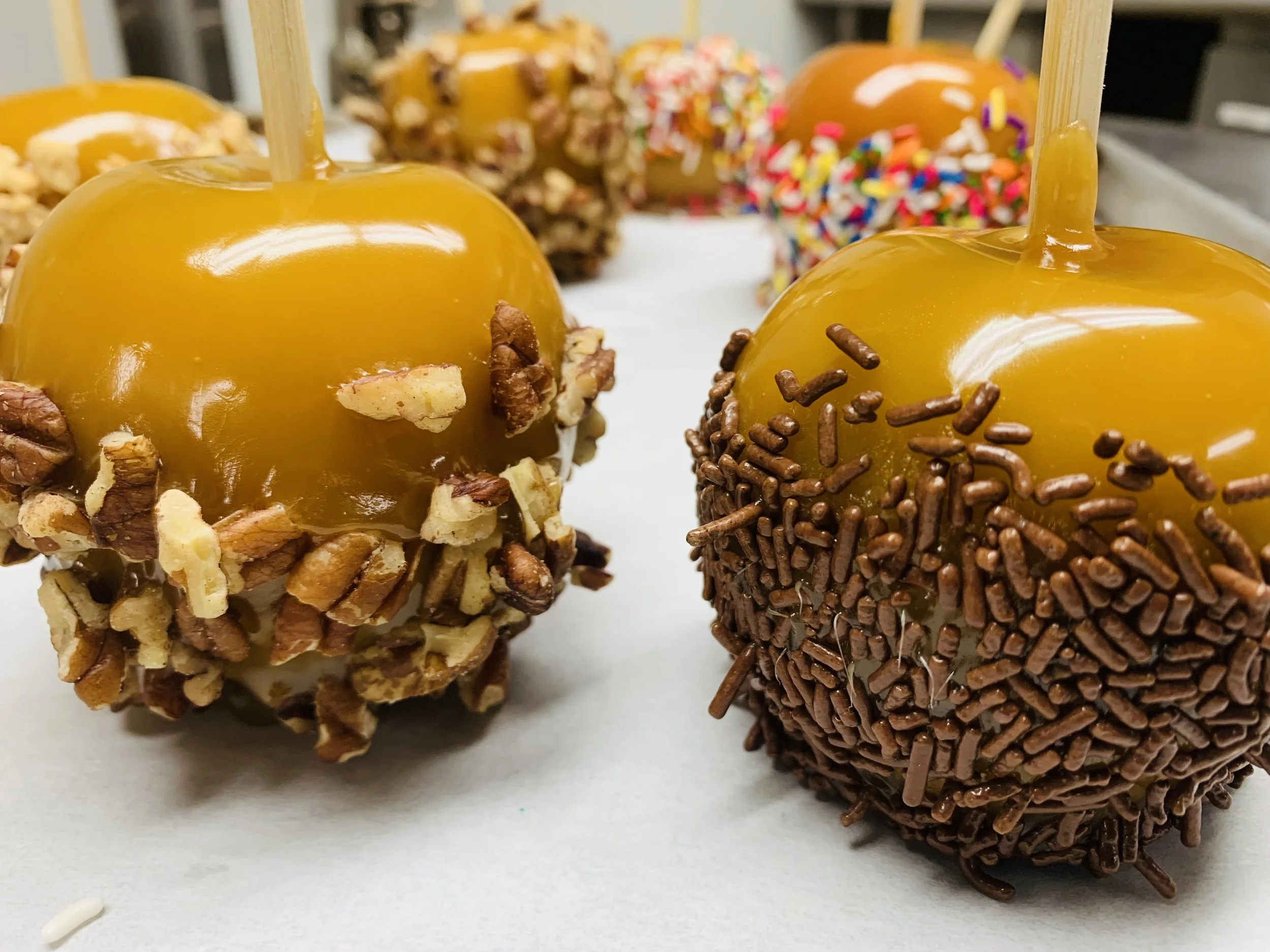 Caramel apples decorated with various toppings such as chopped nuts, colorful sprinkles, and chocolate sprinkles on a white surface.