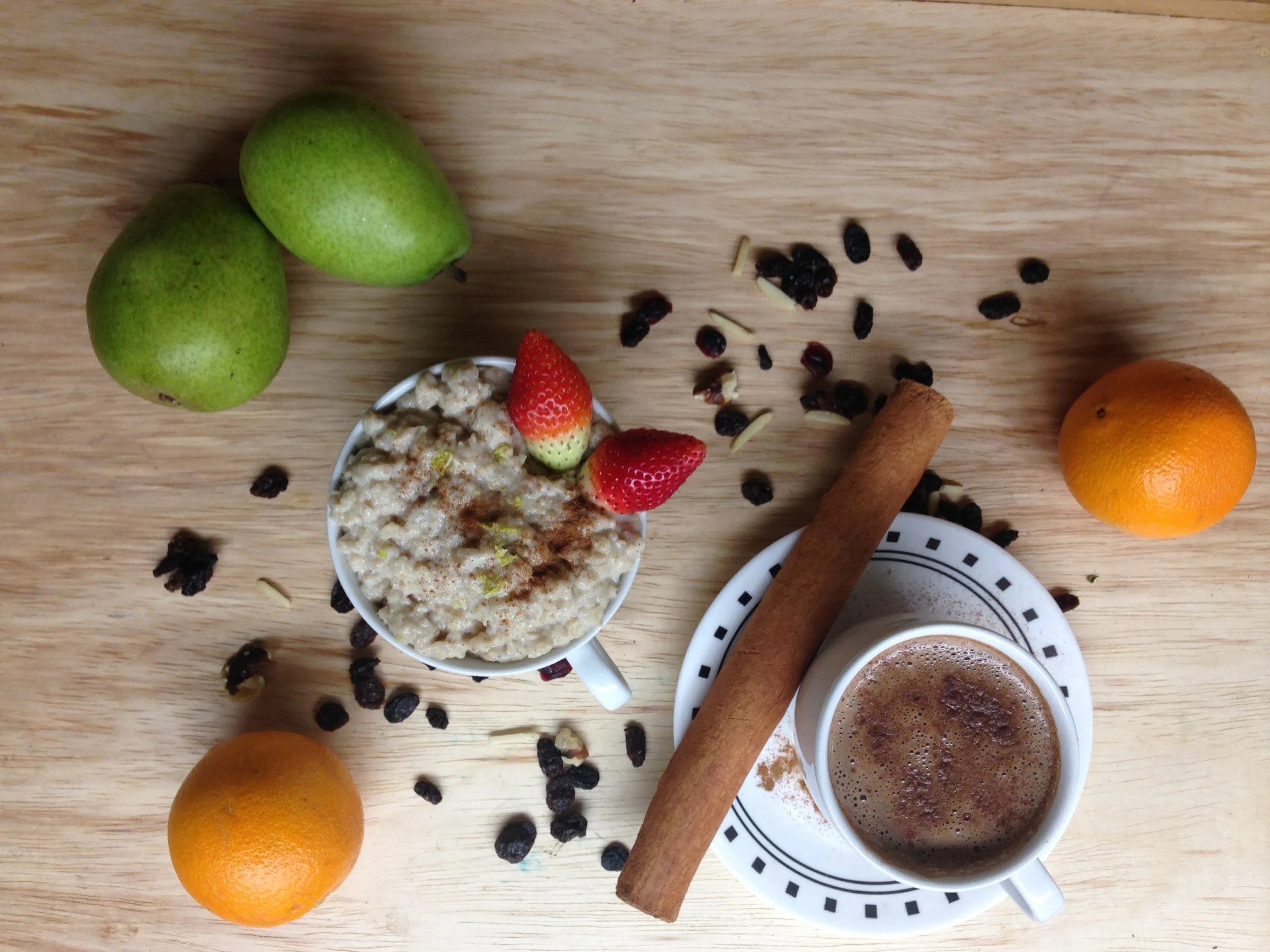 A wooden table containing two green pears, a cup of oatmeal with strawberries, black currants, and nuts, a cinnamon stick, an orange, and two cups of coffee, with scattered black currants and nuts.