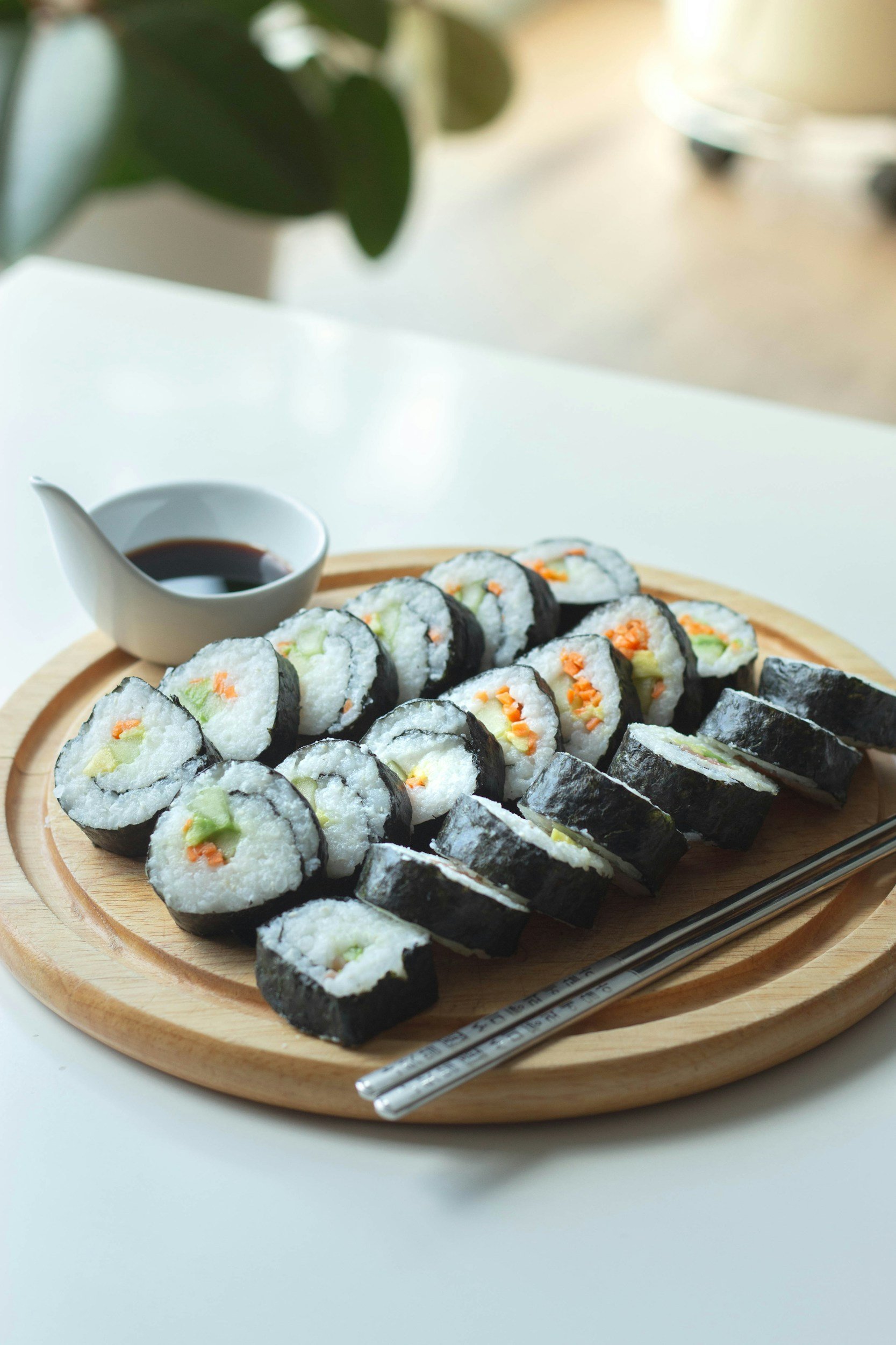 Sushi rolls with vegetables on a wooden tray, soy sauce in a small white dish, chopsticks, and green leaves in the background.