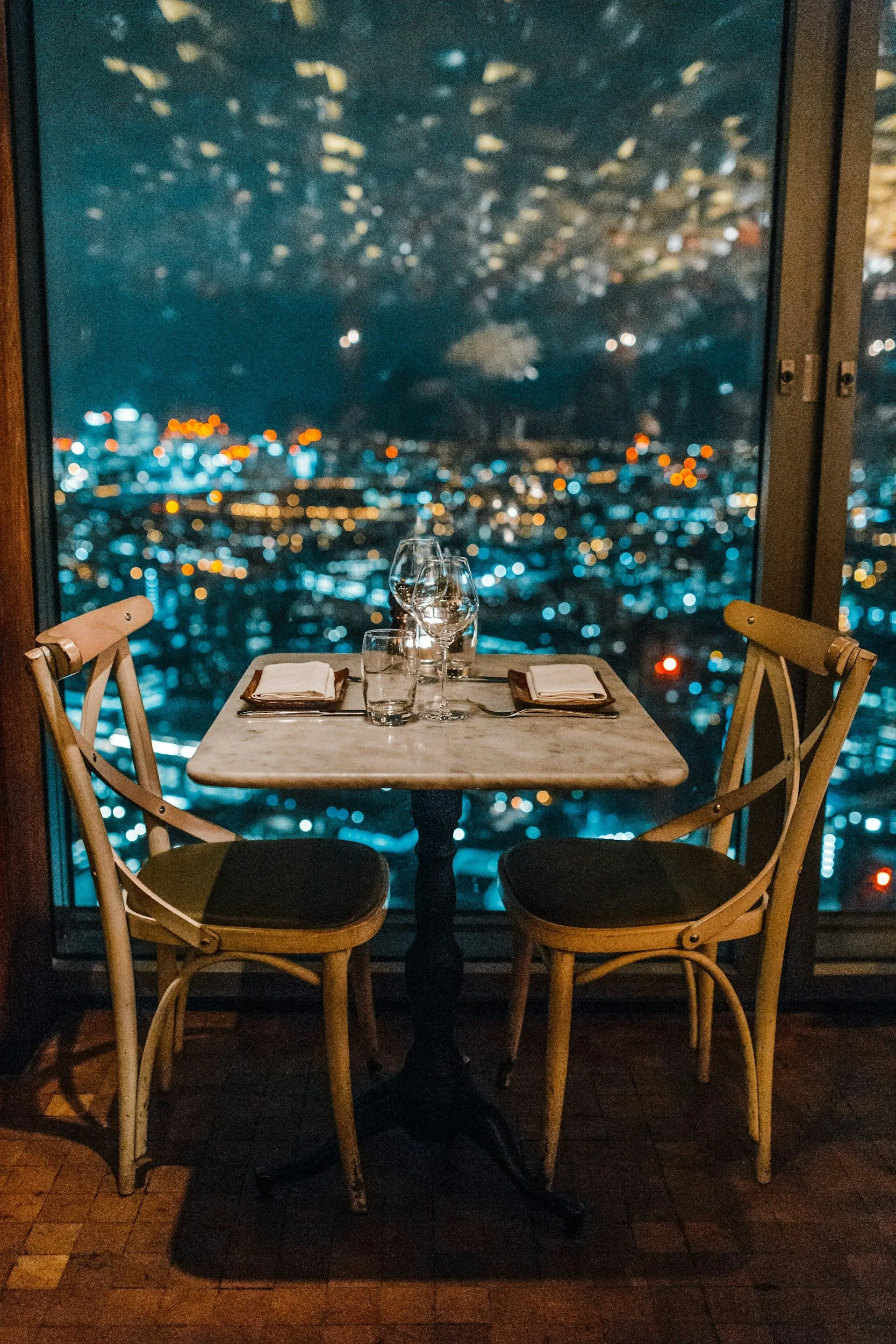 A table set for two with two wine glasses, two water glasses, and two napkins in a restaurant with a large window overlooking a city skyline at night, with blurred city lights and a starry sky.